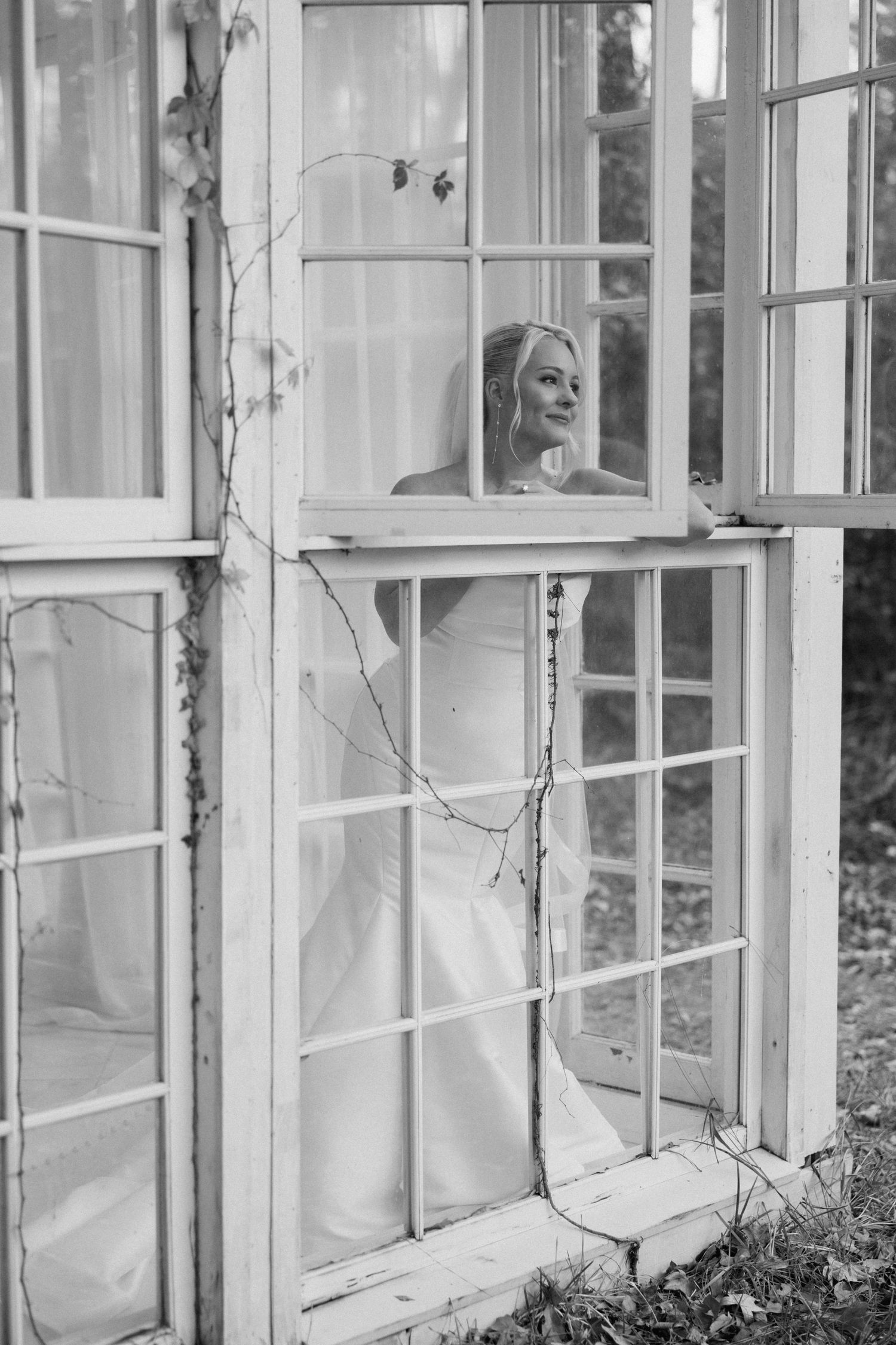 Black and white photo of a bride in a white gown standing inside a vintage greenhouse with white-framed windows.