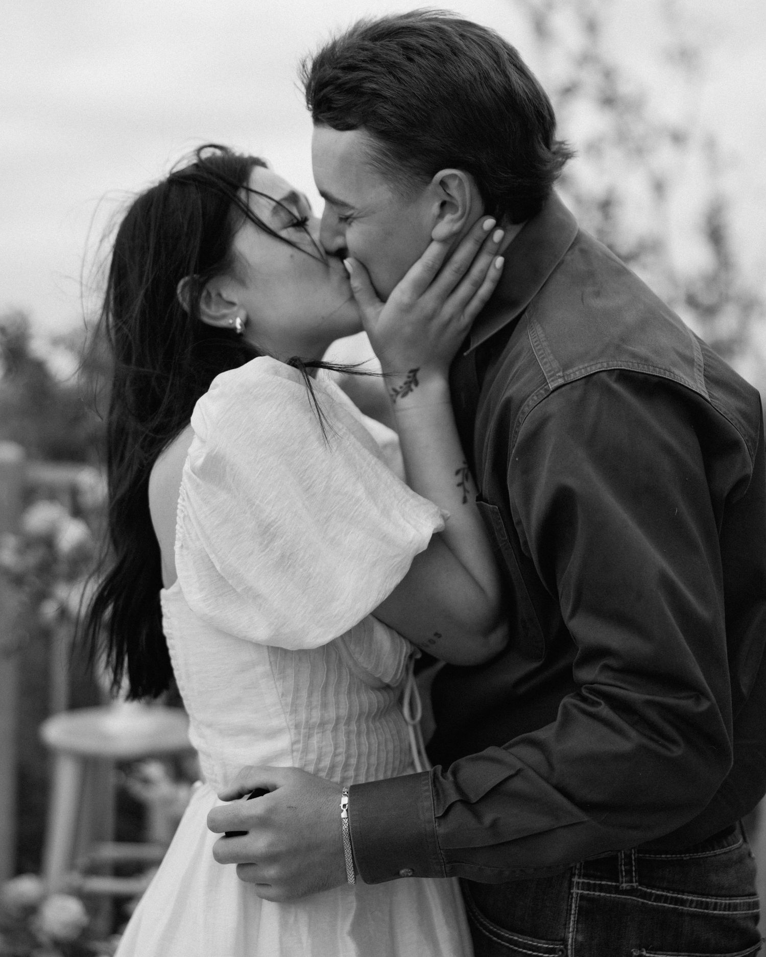 Black and white photo of a couple sharing a romantic kiss outdoors, woman in white dress.