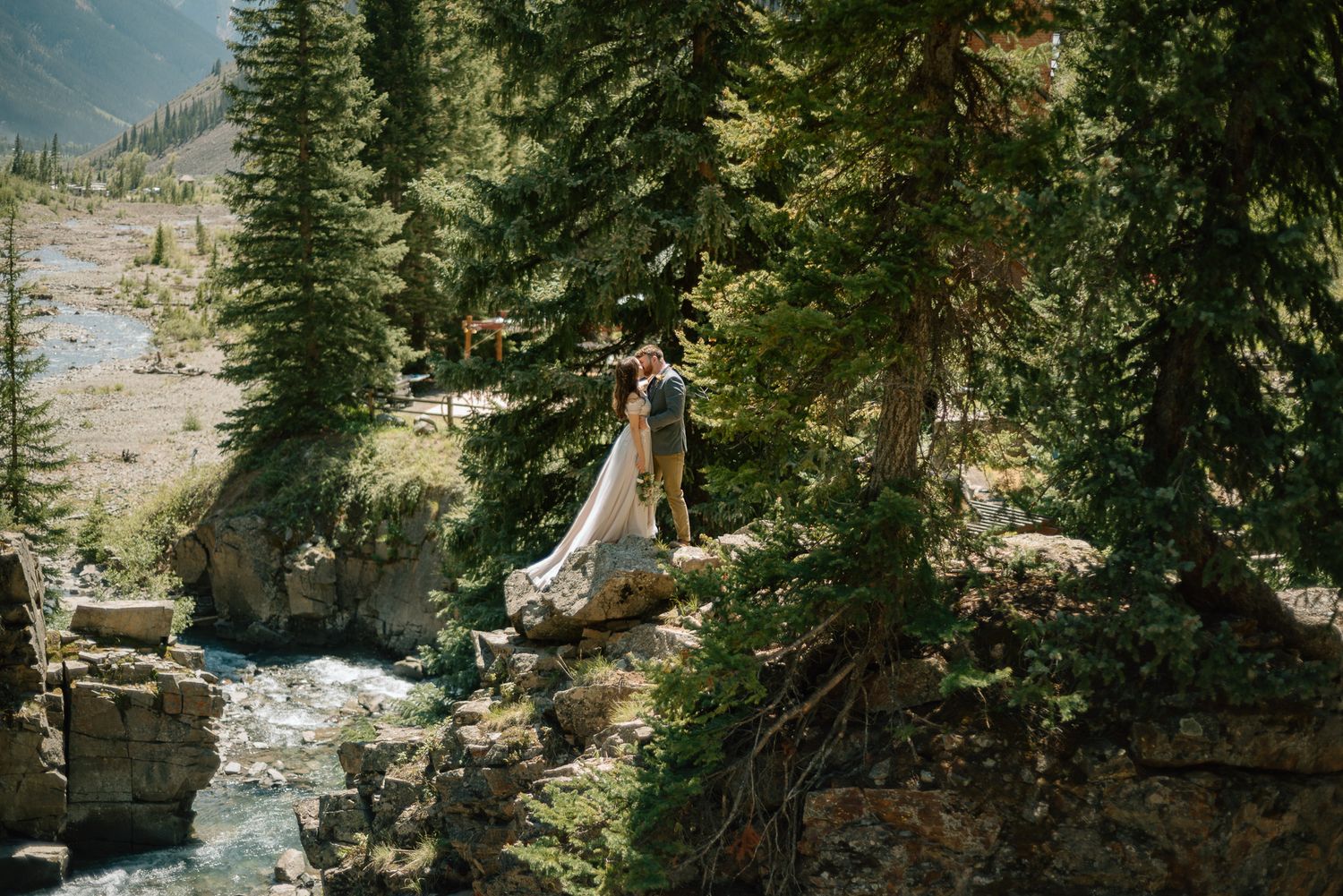 Scenic mountain wedding photo with couple on stone cliff overlooking rushing stream below.