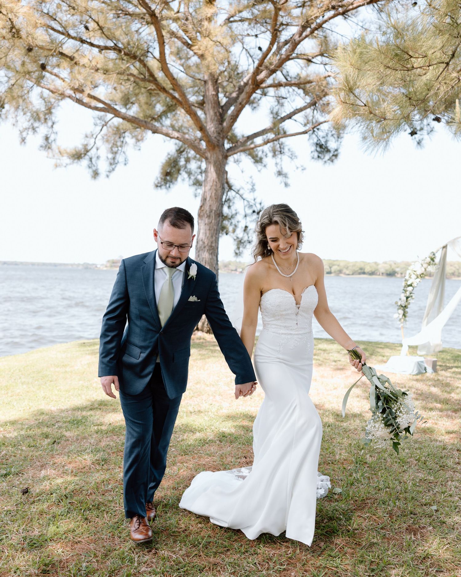 A couple walking hand in hand by a waterfront, with the bride in a fitted white dress and groom in a navy suit.