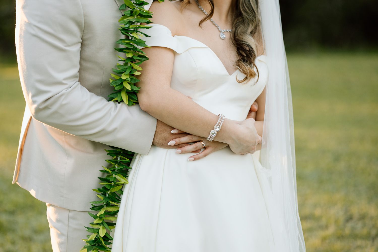 Close up detail of bride and groom embracing, showing off the maile lei and white wedding dress.