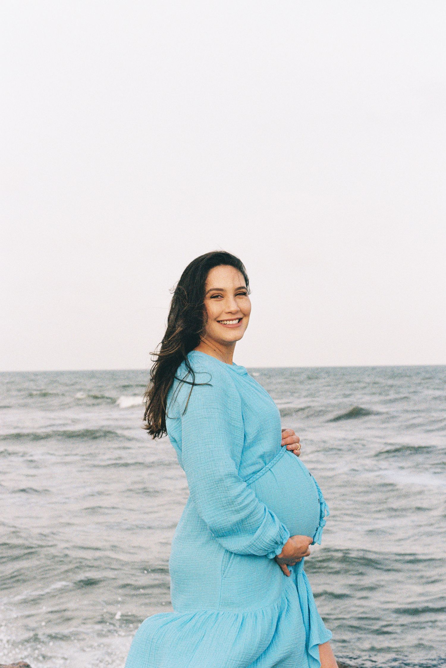 A pregnant woman in a blue dress poses by the ocean with a joyful expression.