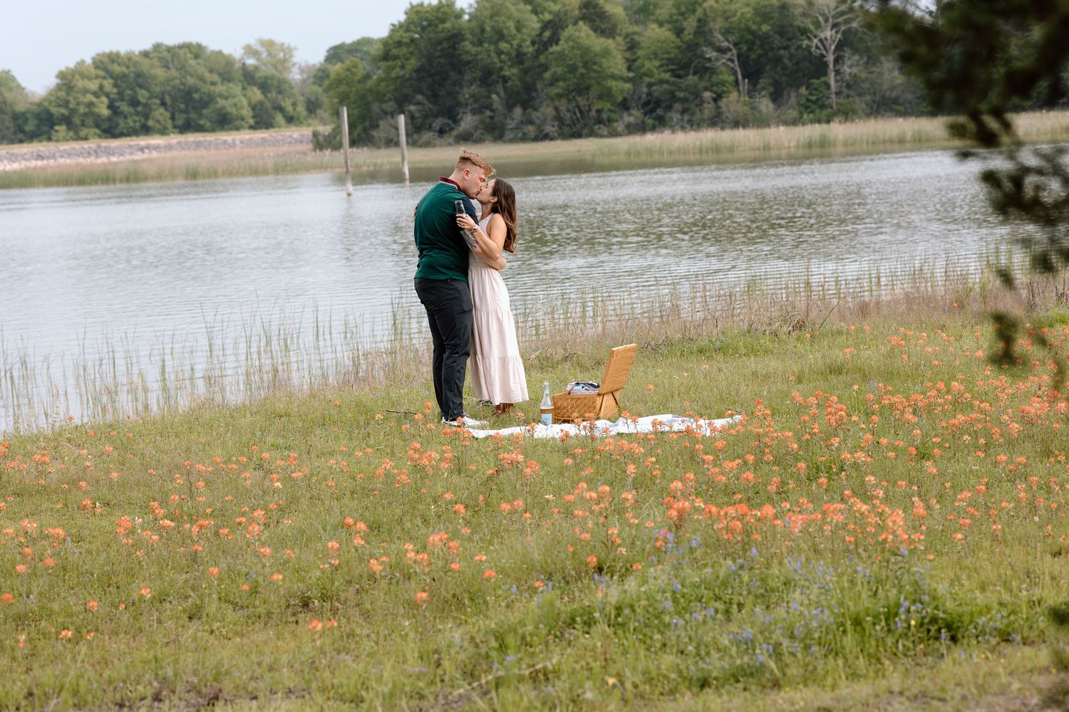 A couple stands together on a lakeside field surrounded by orange wildflowers and a picnic setup.