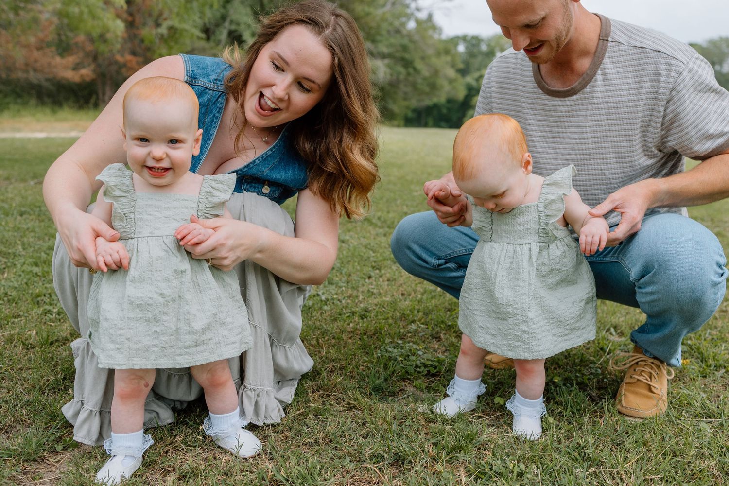 Two babies in matching gray dresses smile with their parents while sitting on green grass in a park setting.