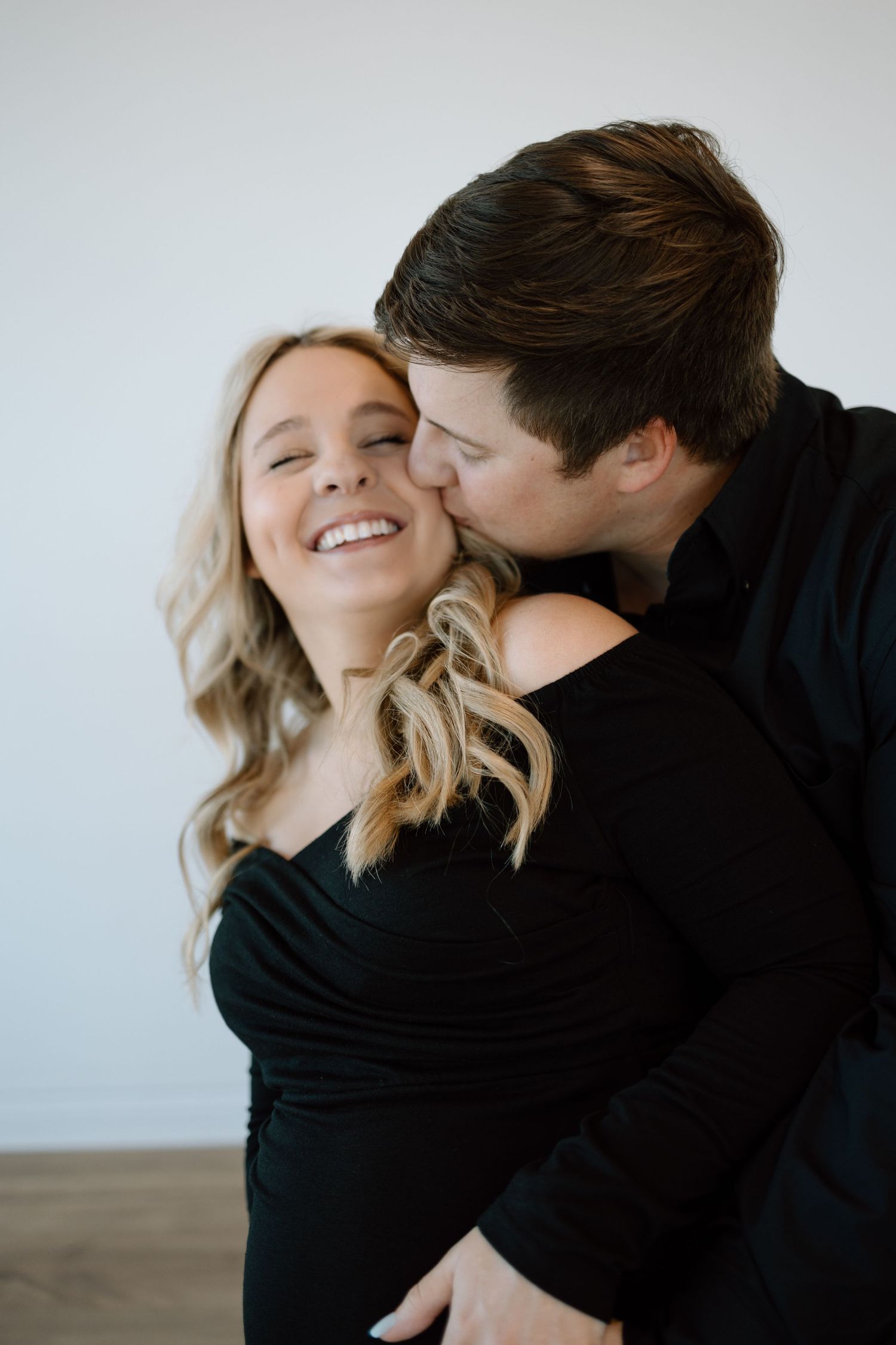 A loving couple shares an intimate moment as they embrace against a white wall, with the woman wearing black.