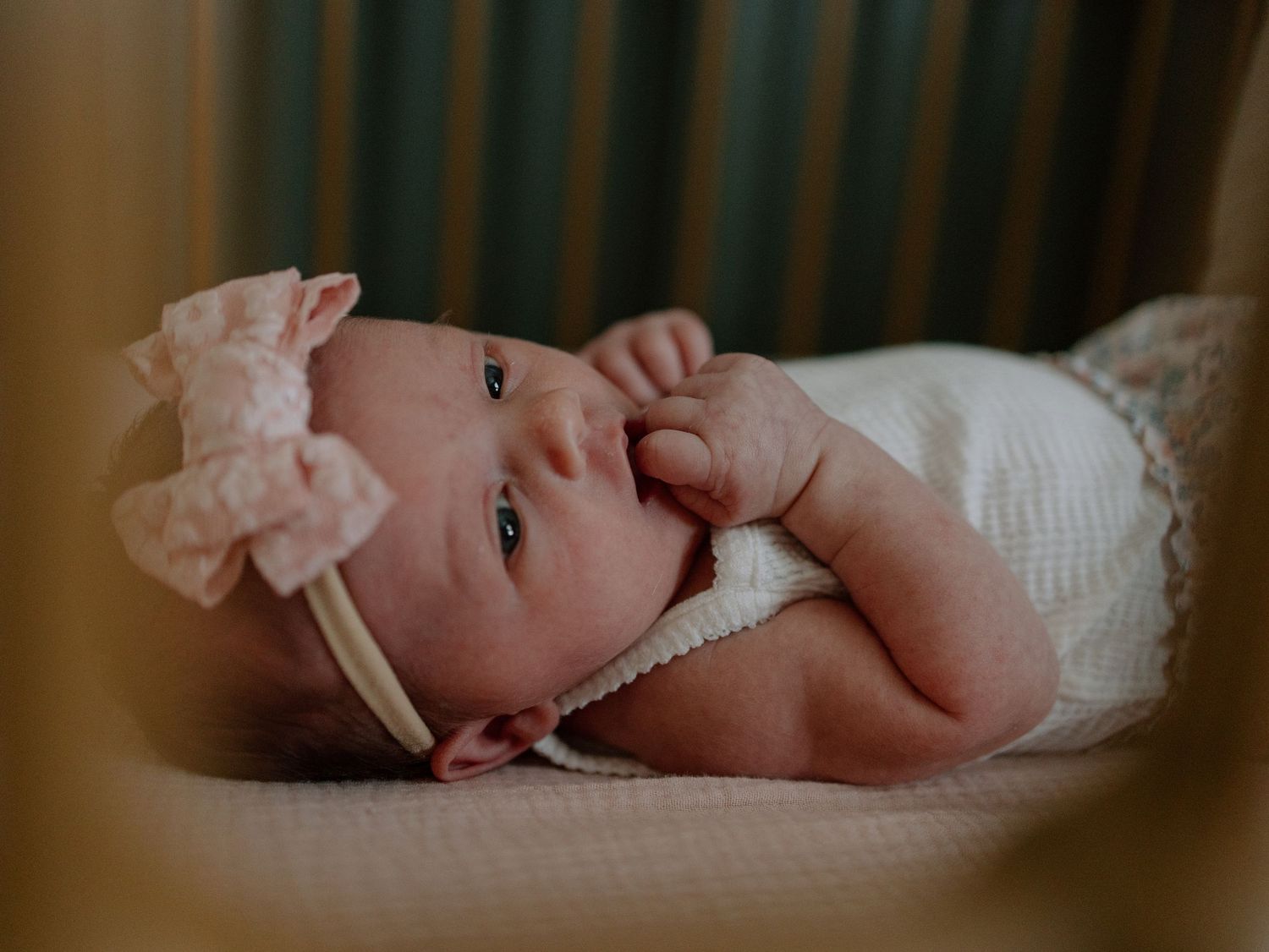 Baby wearing white headband and outfit lying down with hand near mouth in soft lighting.