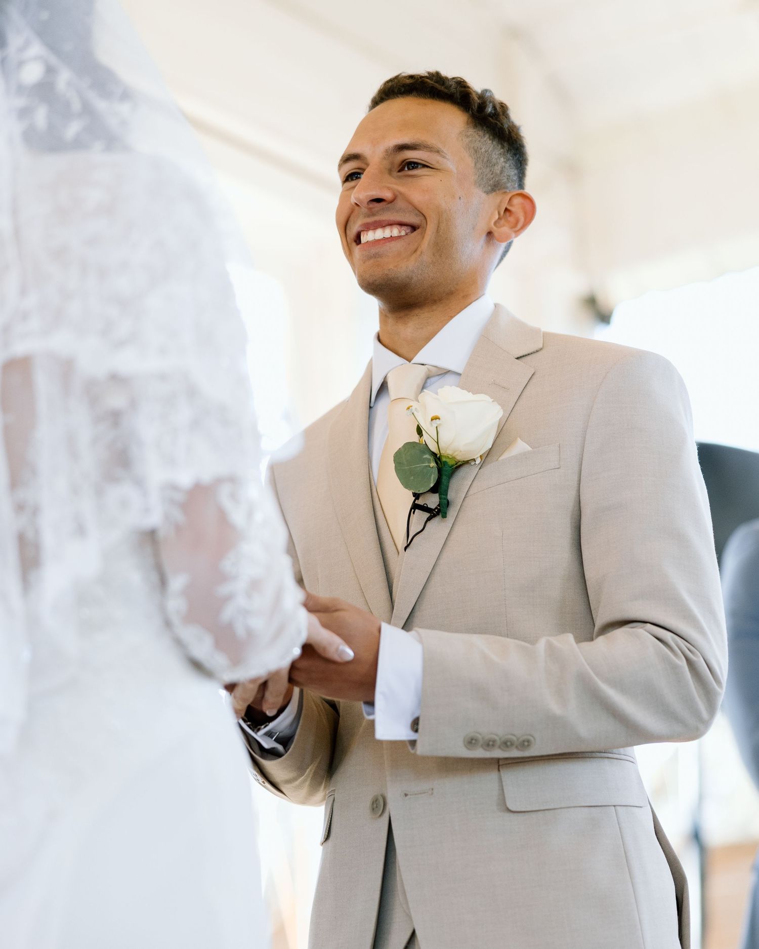 Groom in tan suit smiling joyfully as bride approaches during wedding ceremony.