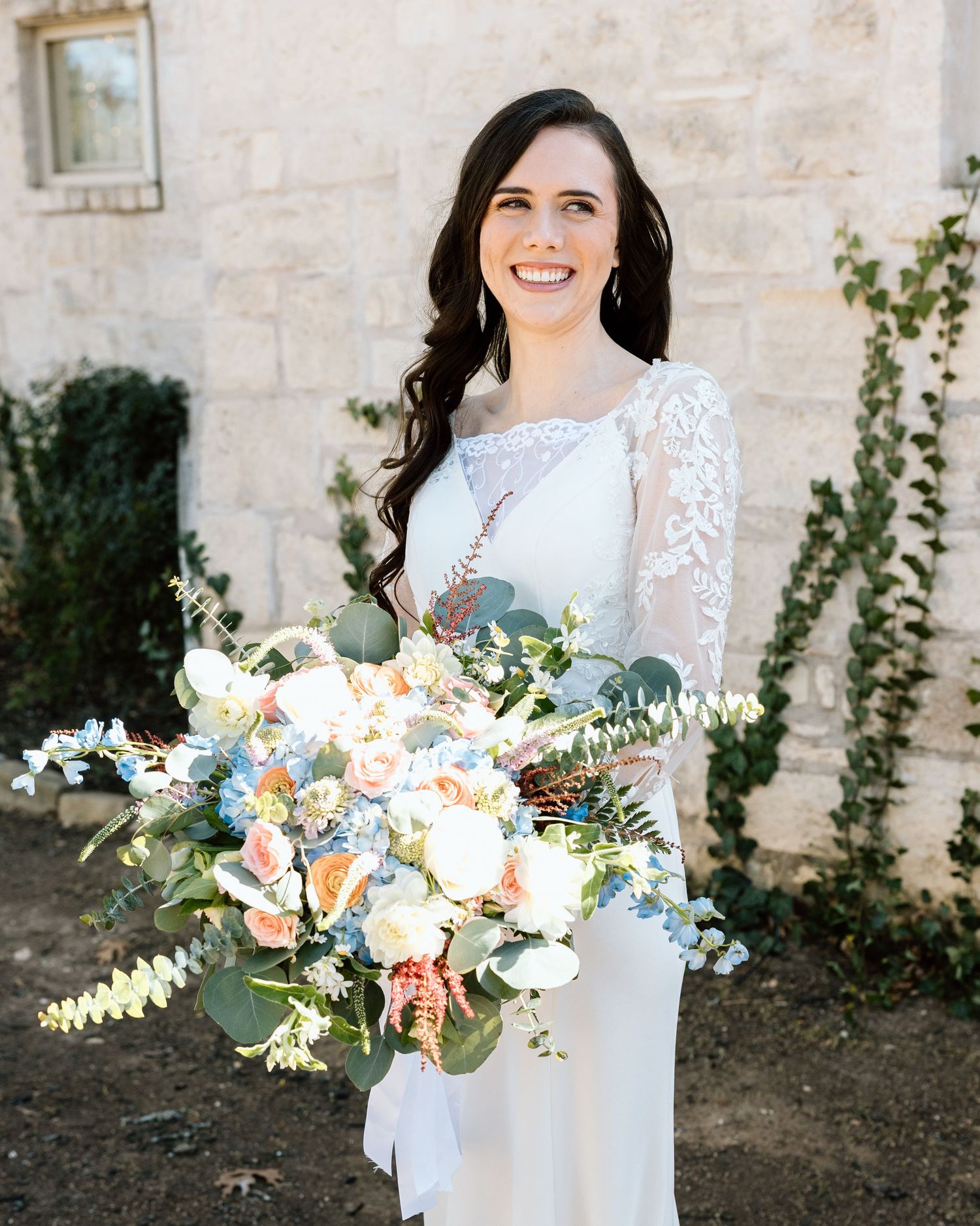 Smiling bride in white lace gown holding large bouquet of peach, white, and blue flowers with eucalyptus greenery.