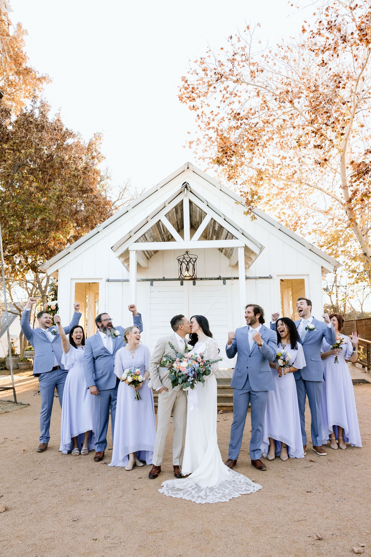 Wedding party in blue suits and lavender gowns cheering as bride and groom kiss before white barn chapel.