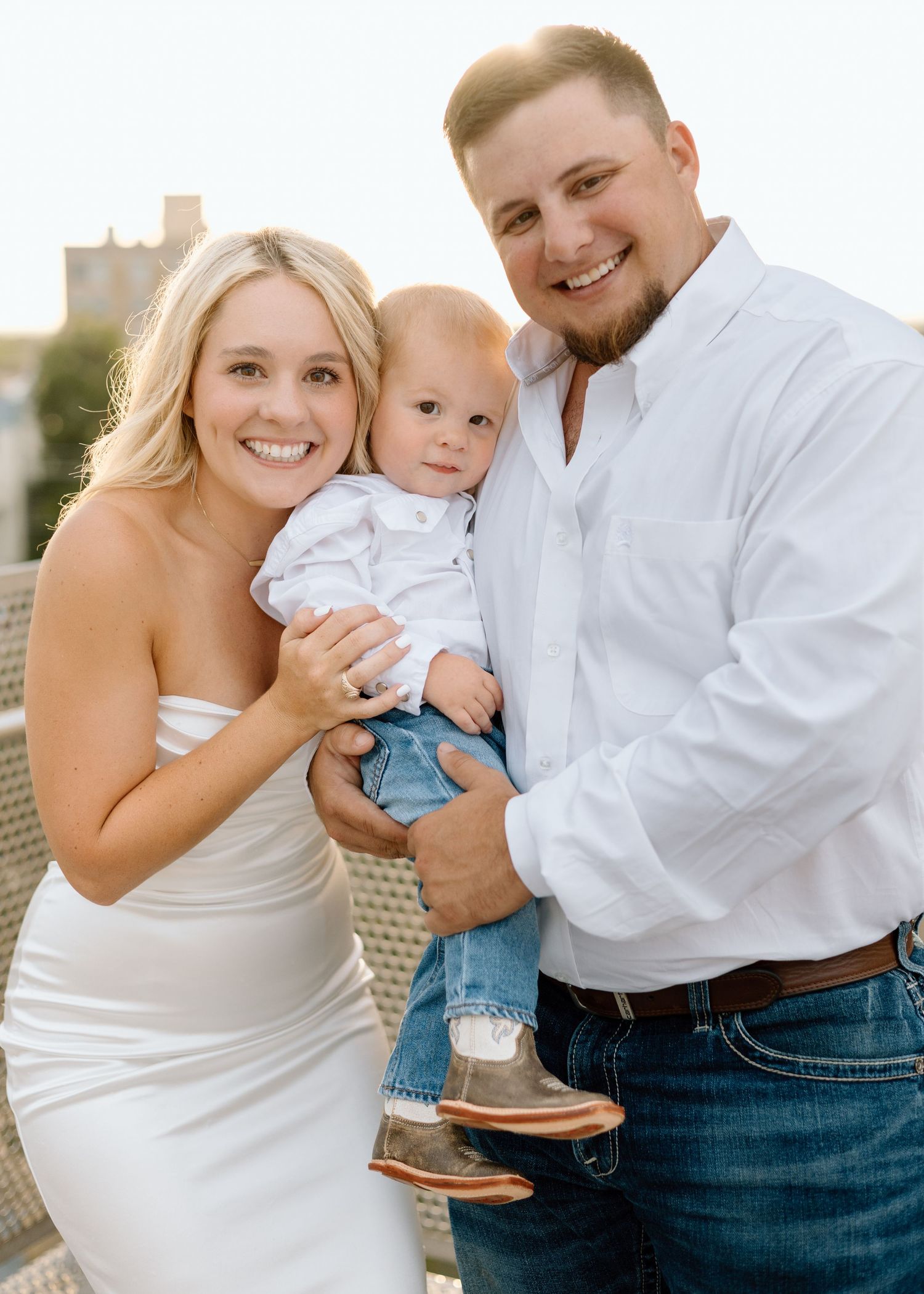 A smiling couple in white outfits holds their baby boy outdoors during a golden hour family portrait session.