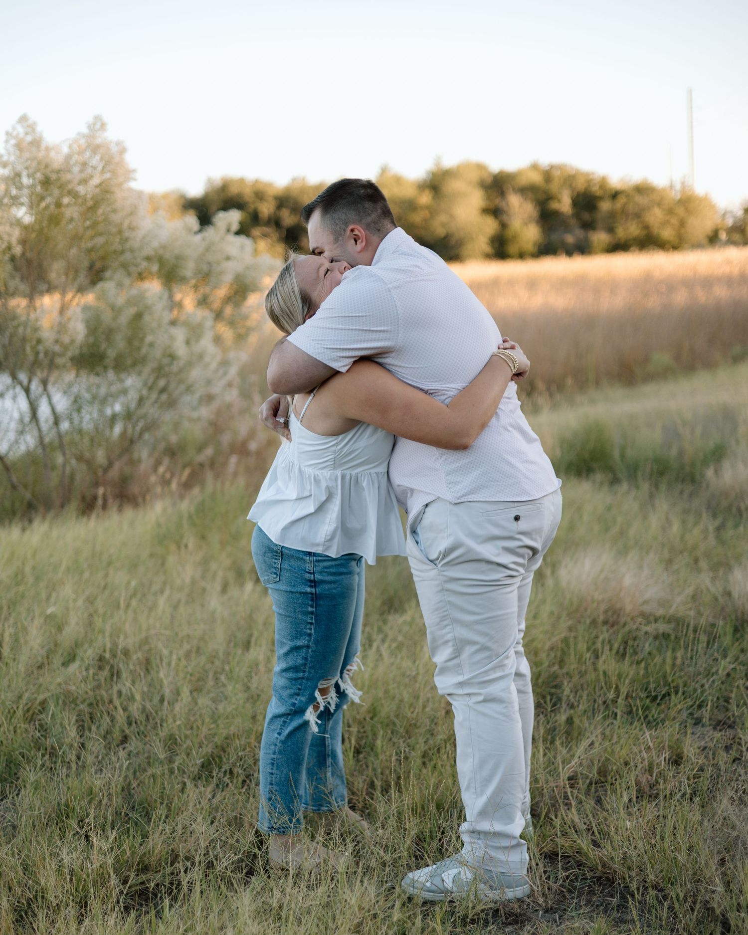 Couple embracing and kissing in a golden field at sunset, wearing white tops and jeans outdoors.