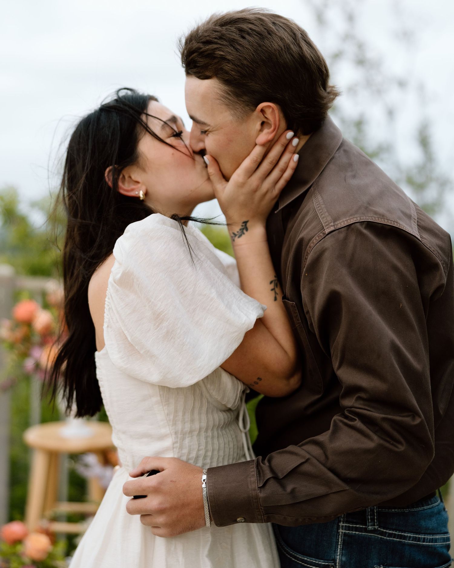 A couple shares a romantic kiss outdoors, with the woman in a white dress and man in a brown leather jacket.