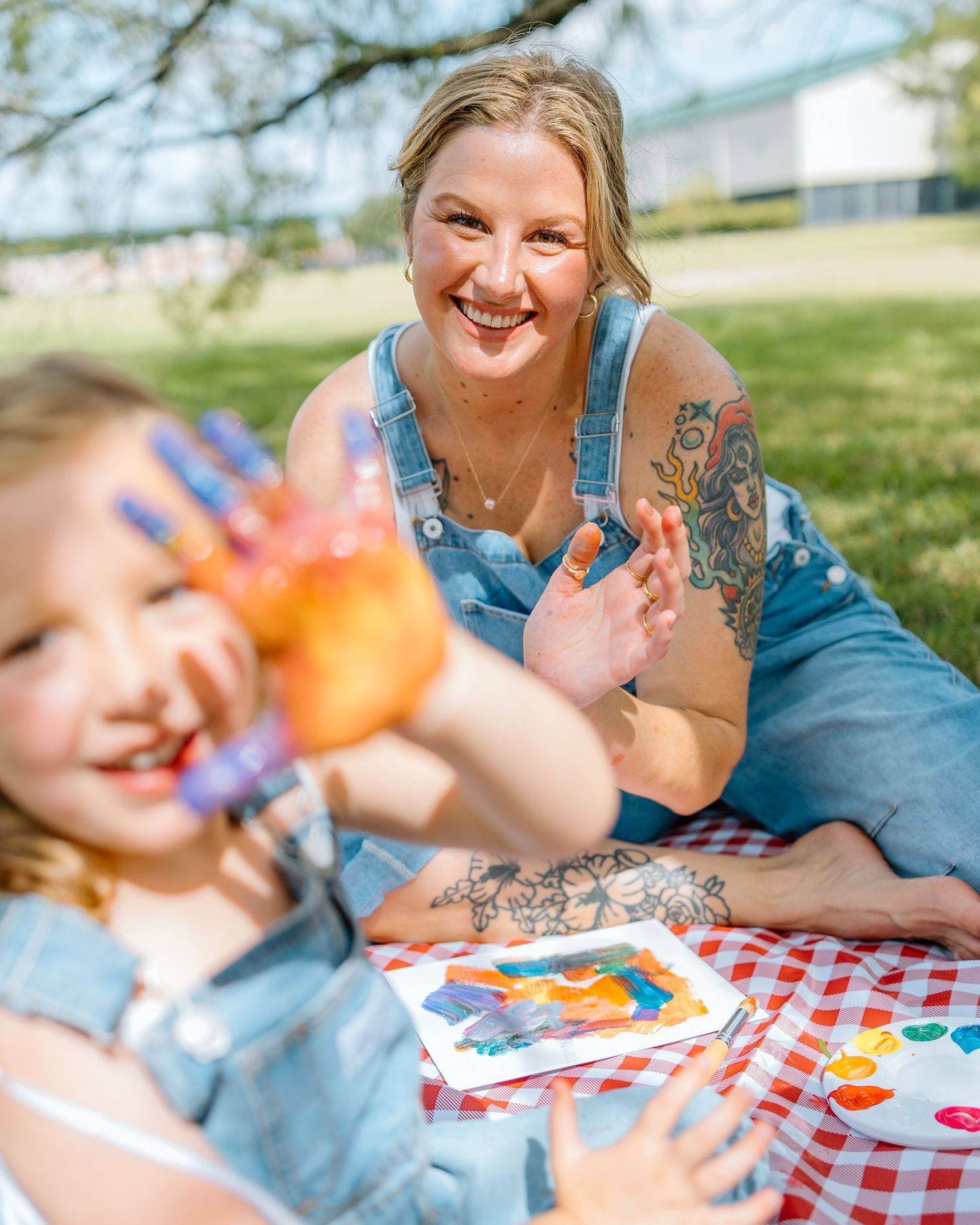 Woman and young girl doing colorful finger painting together on a picnic blanket outdoors in sunny park.