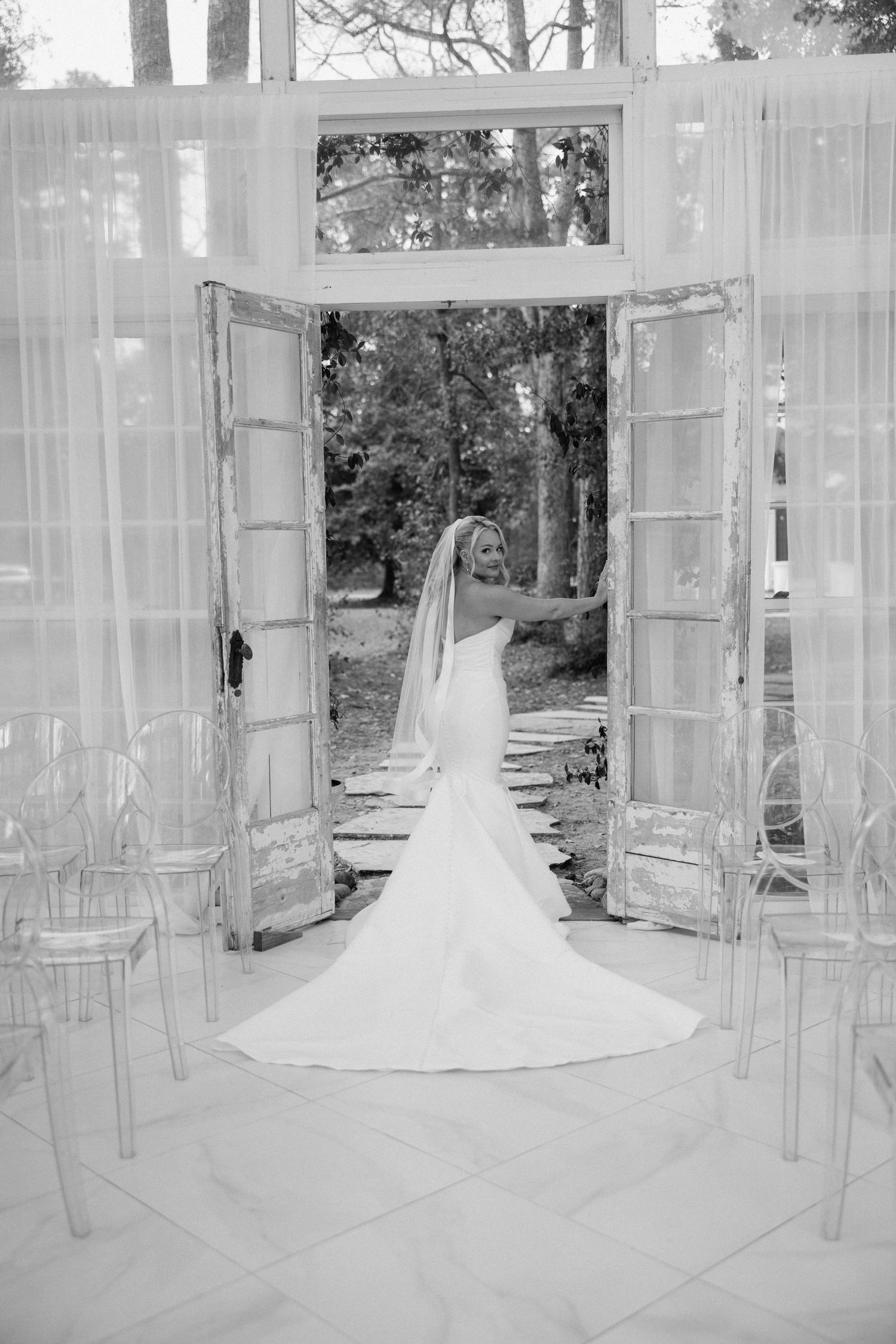 Bride in elegant mermaid wedding gown standing in doorway of white venue in black and white photo.