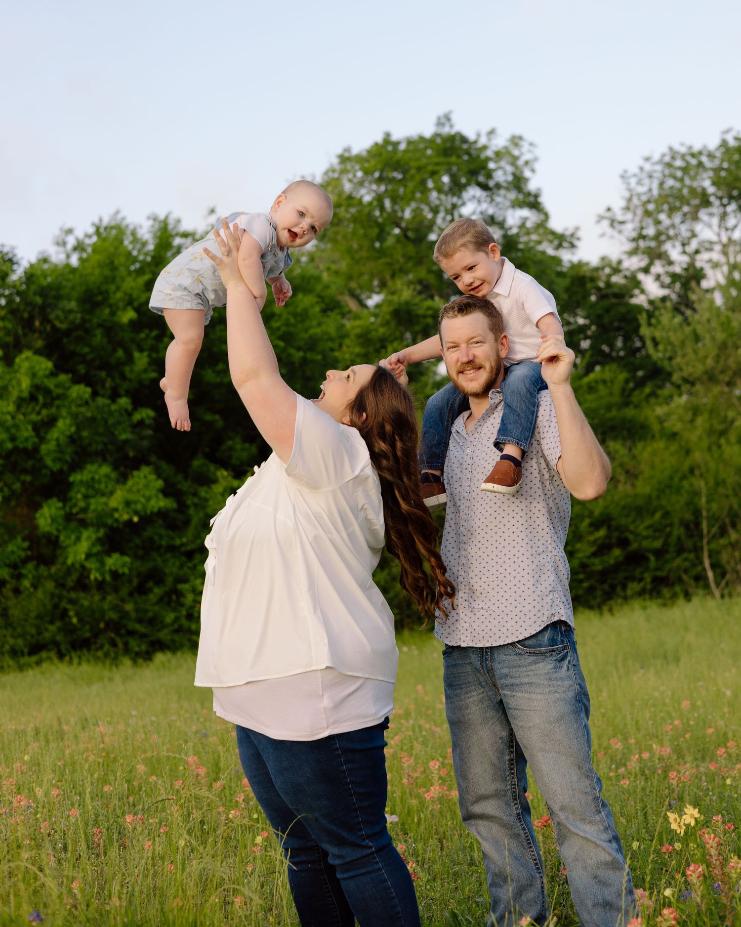 Two adults lift small children onto their shoulders while standing together in a grassy meadow with trees in the background.