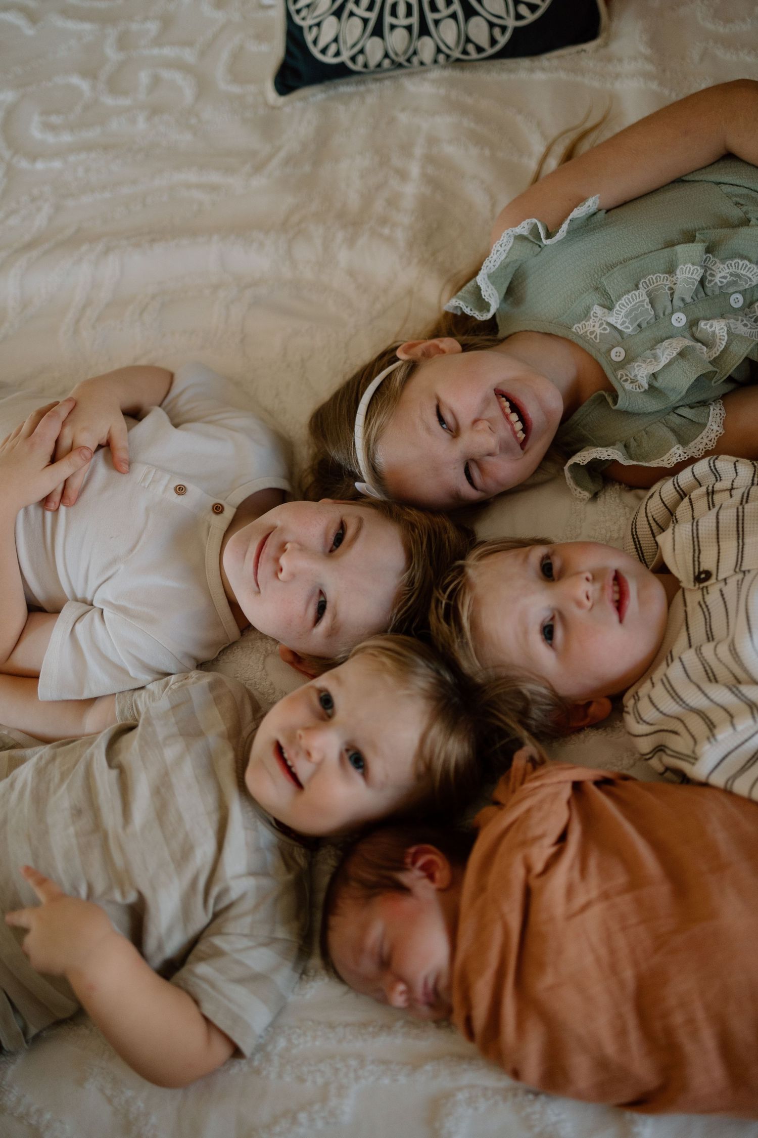 A group of five young siblings lying together in a circle on a bed, wearing casual clothing in earth tones.