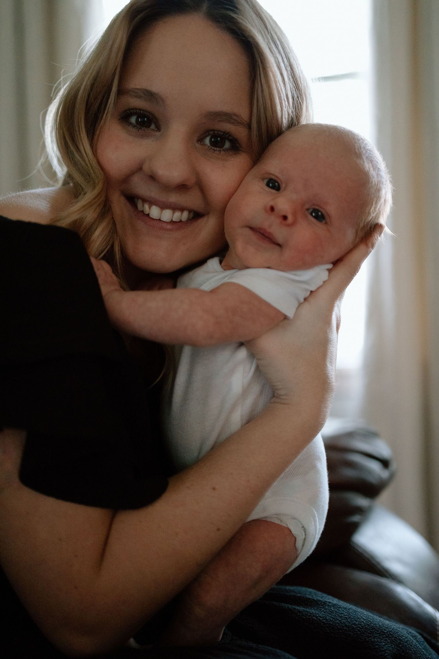 A joyful moment shared between someone wearing black and their young infant as they pose together on a couch.