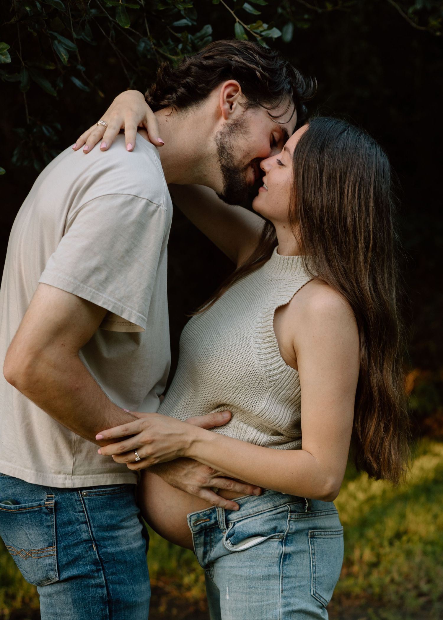 Intimate maternity photo of couple in casual wear against garden background.