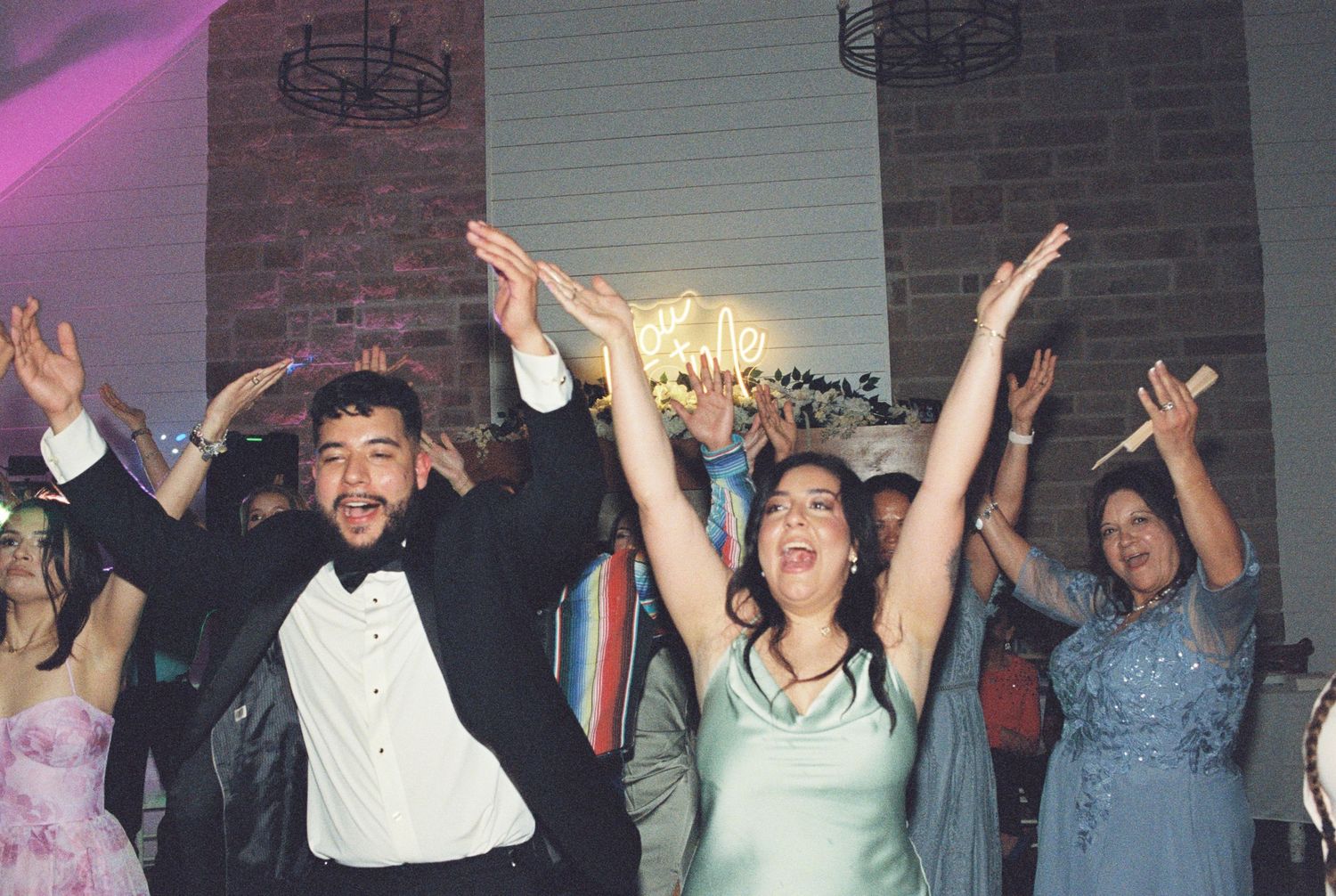 Enthusiastic wedding guests dancing with arms raised during reception celebration.