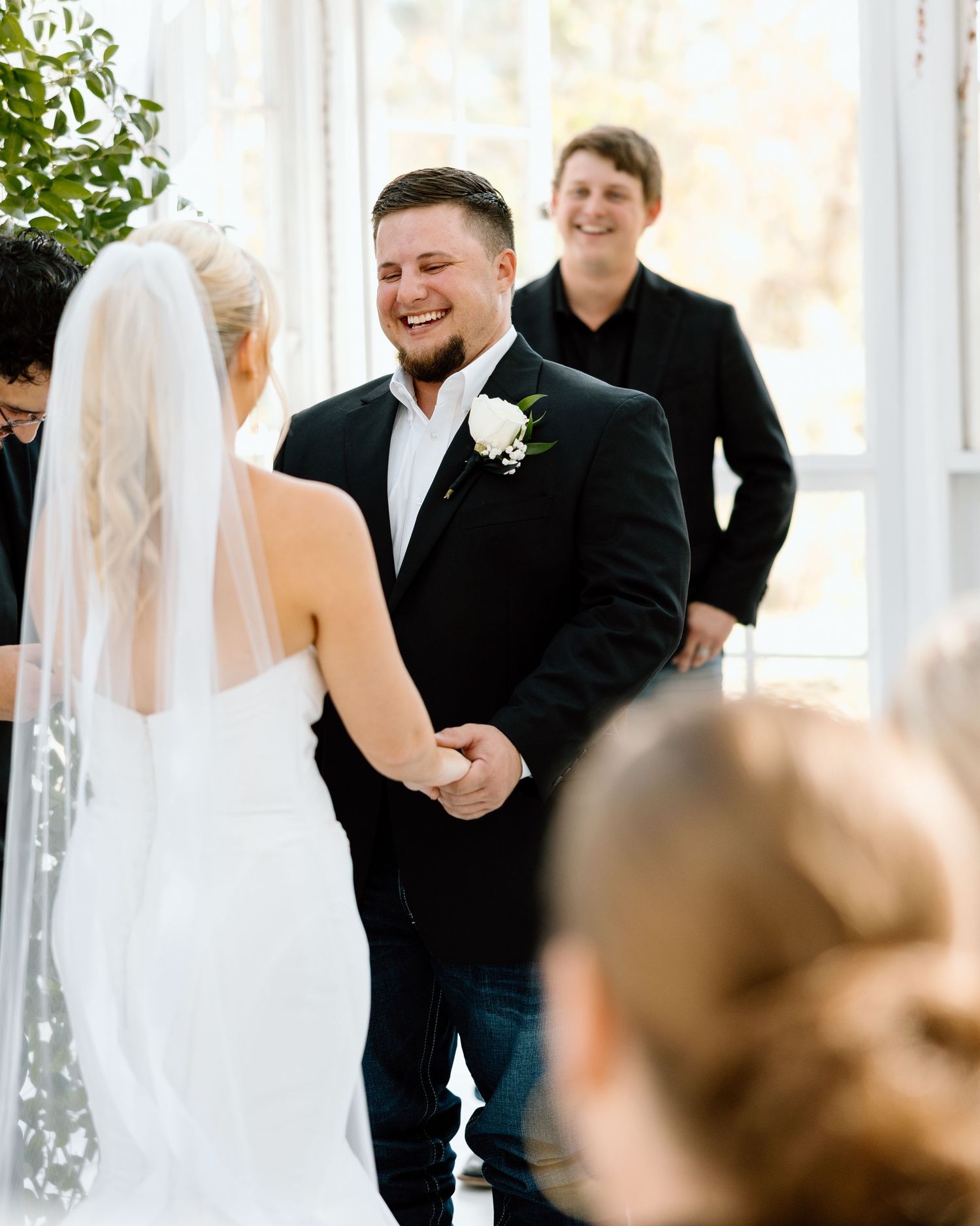 Groom laughing joyfully while holding hands with bride in white gown and veil during indoor wedding ceremony.
