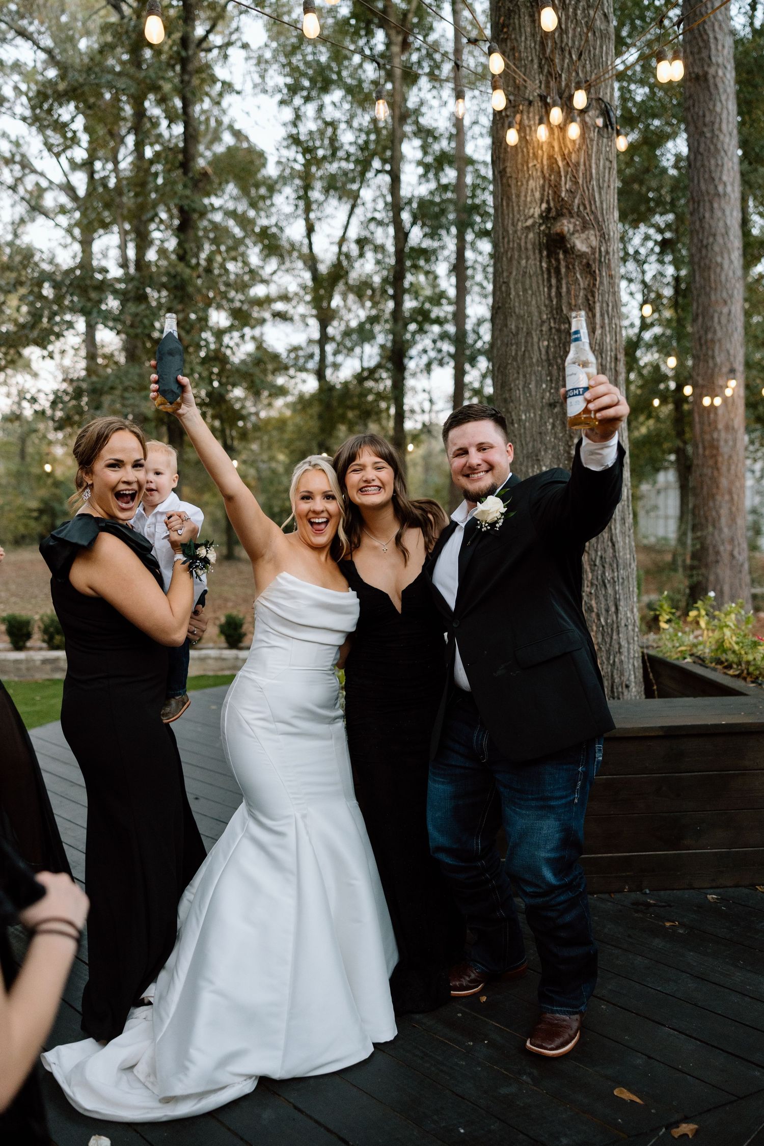 Bride and groom celebrate with wedding party guests holding drinks and phone under string lights outdoors.