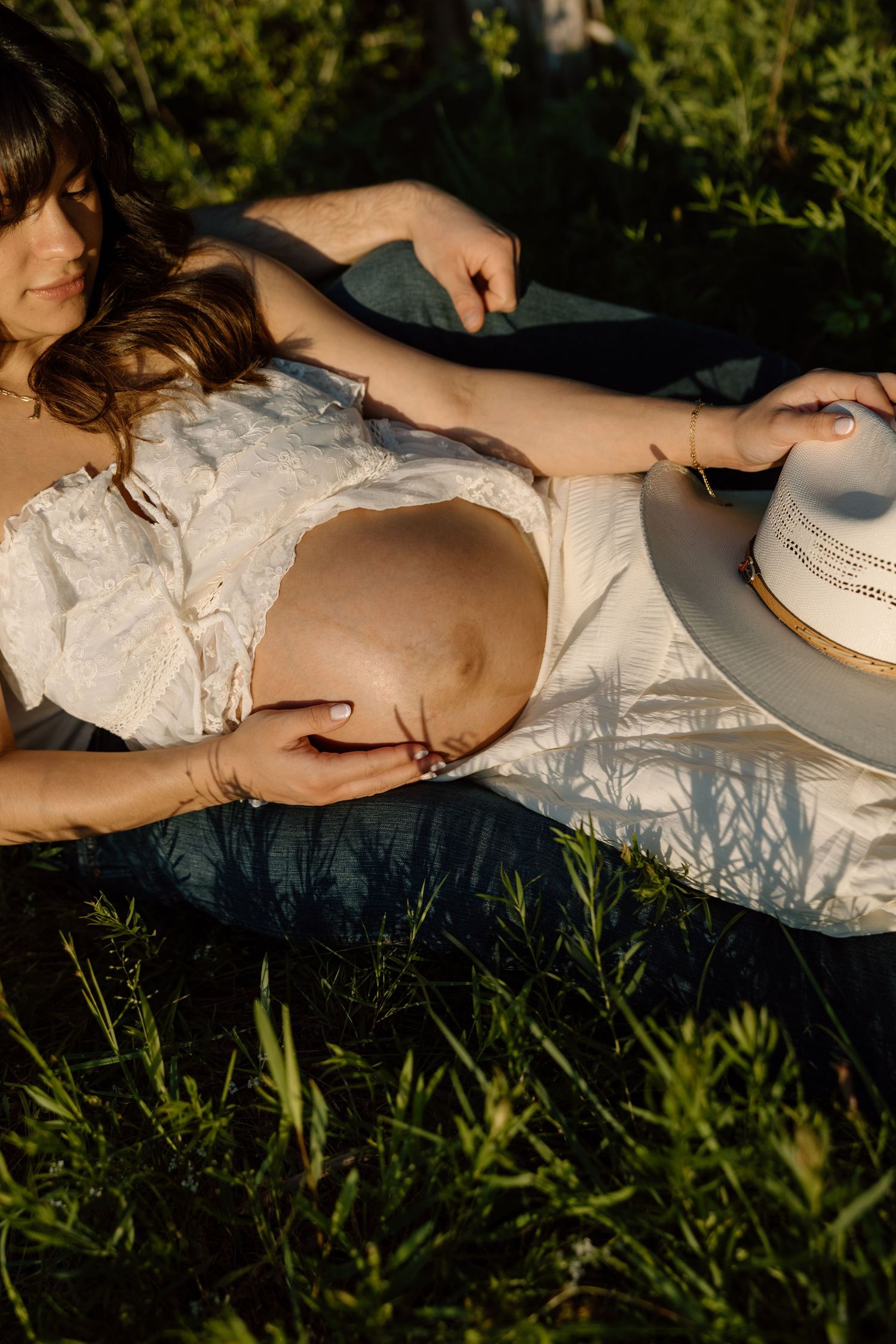 Pregnant woman in white crop top lying in green grass, cradling baby bump, holding white hat in golden sunlight.
