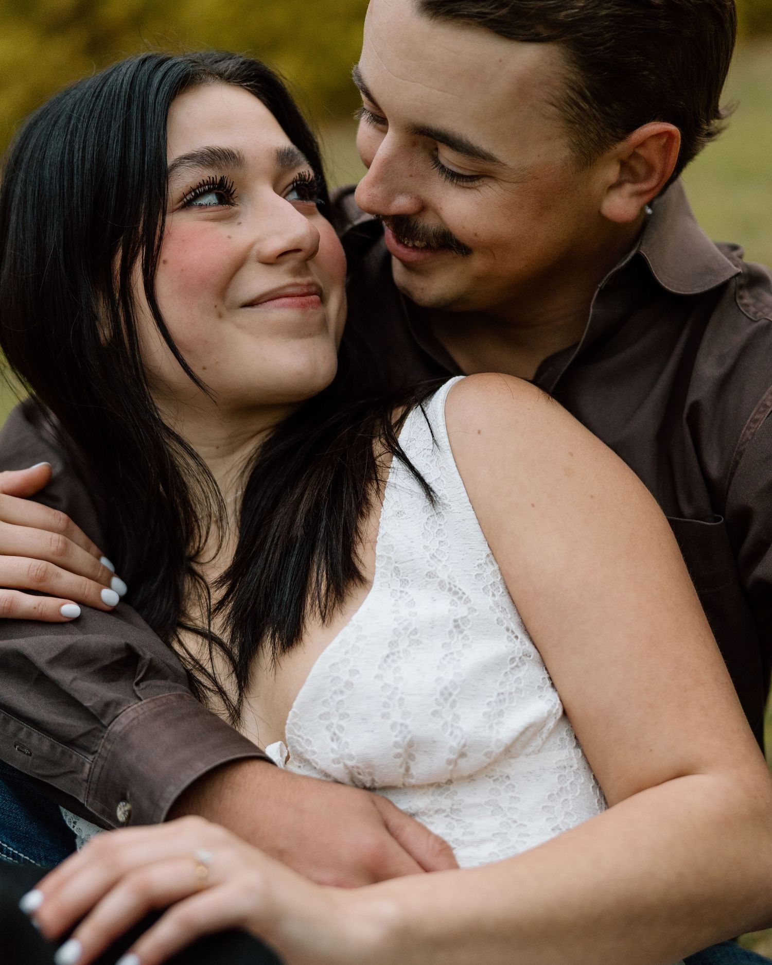 A couple shares a tender embrace outdoors, the woman smiling warmly as the man holds her close among autumn foliage.