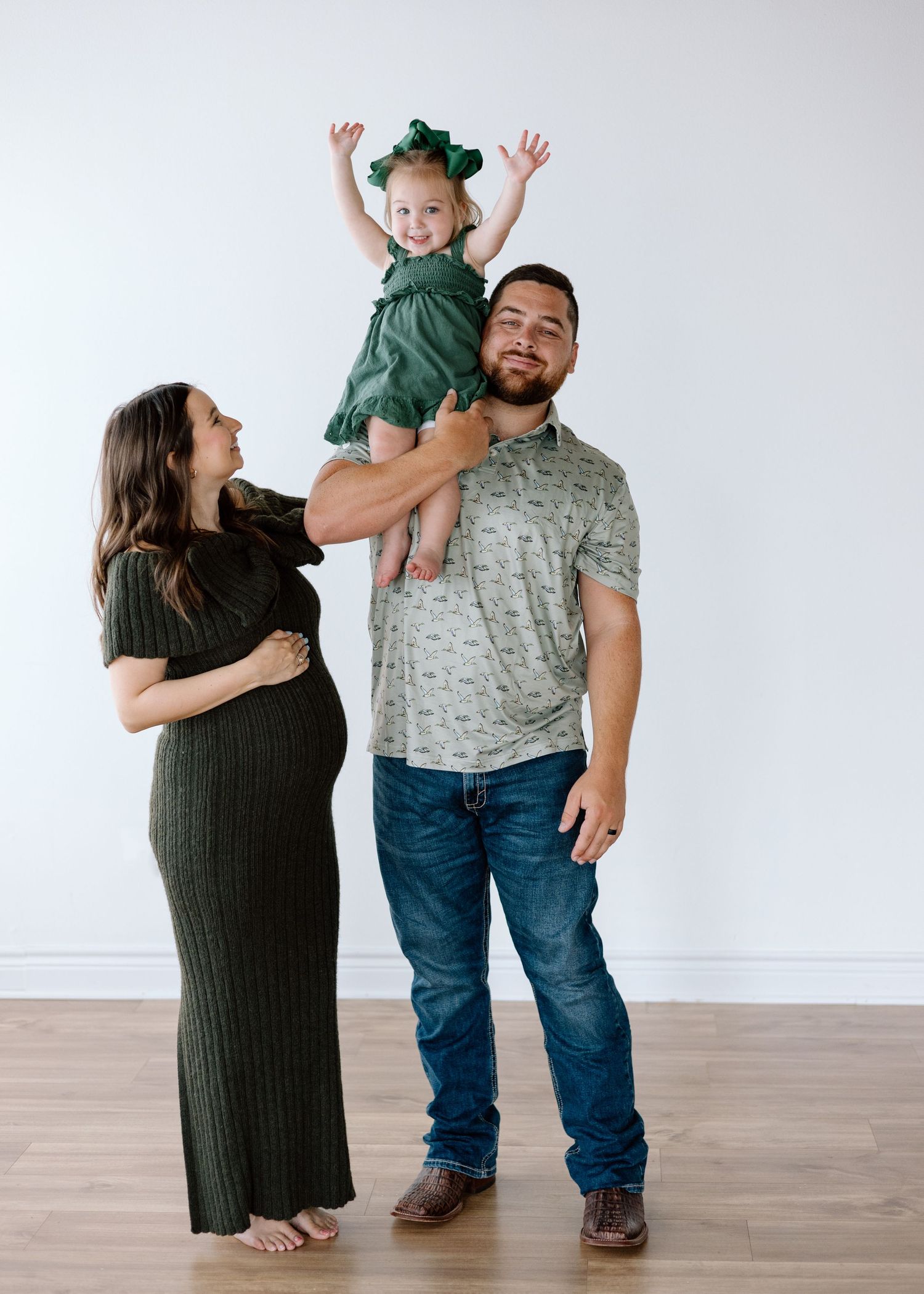 Pregnant woman in green dress with husband holding toddler girl in green outfit, family maternity photo.