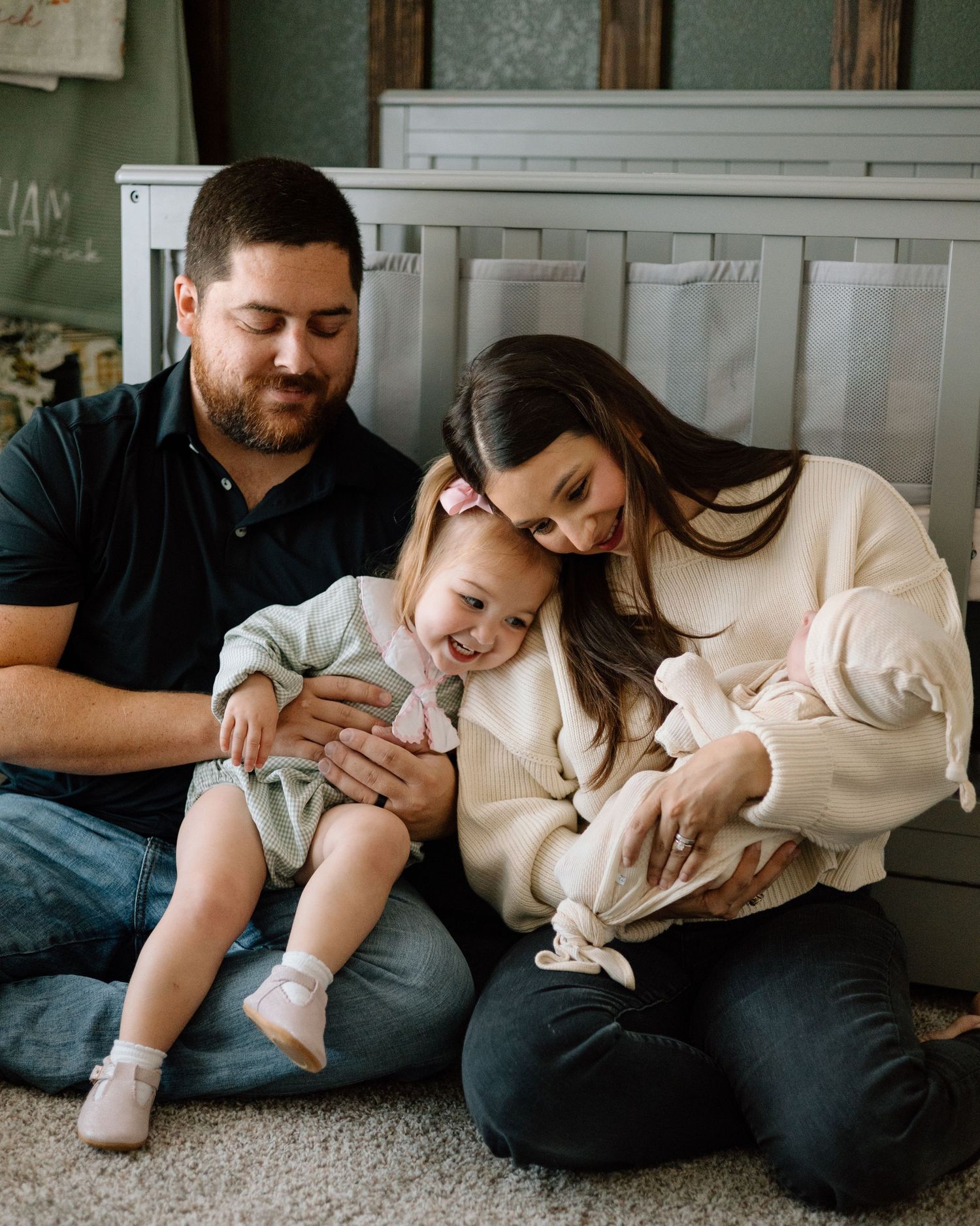 Happy family of four sitting on floor, parents smiling at toddler girl and newborn baby in cozy home.