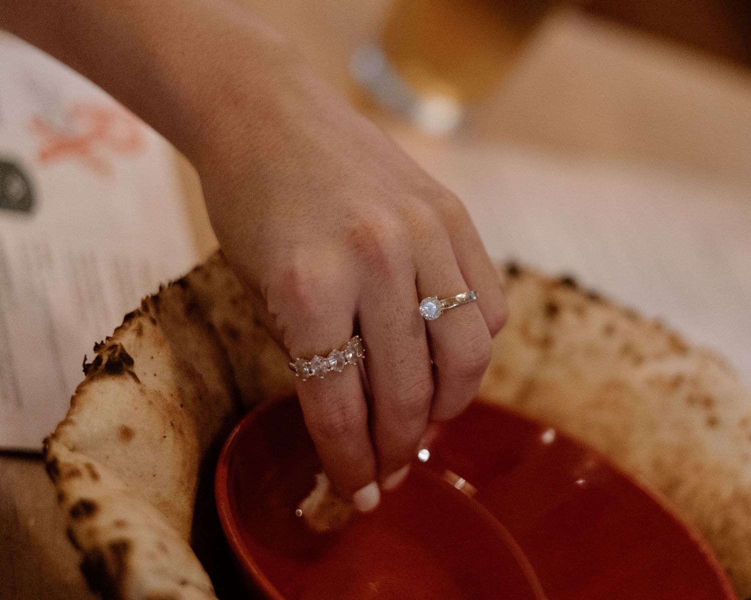 A hand wearing diamond rings reaches toward a red bowl beside golden matzah bread on a table.