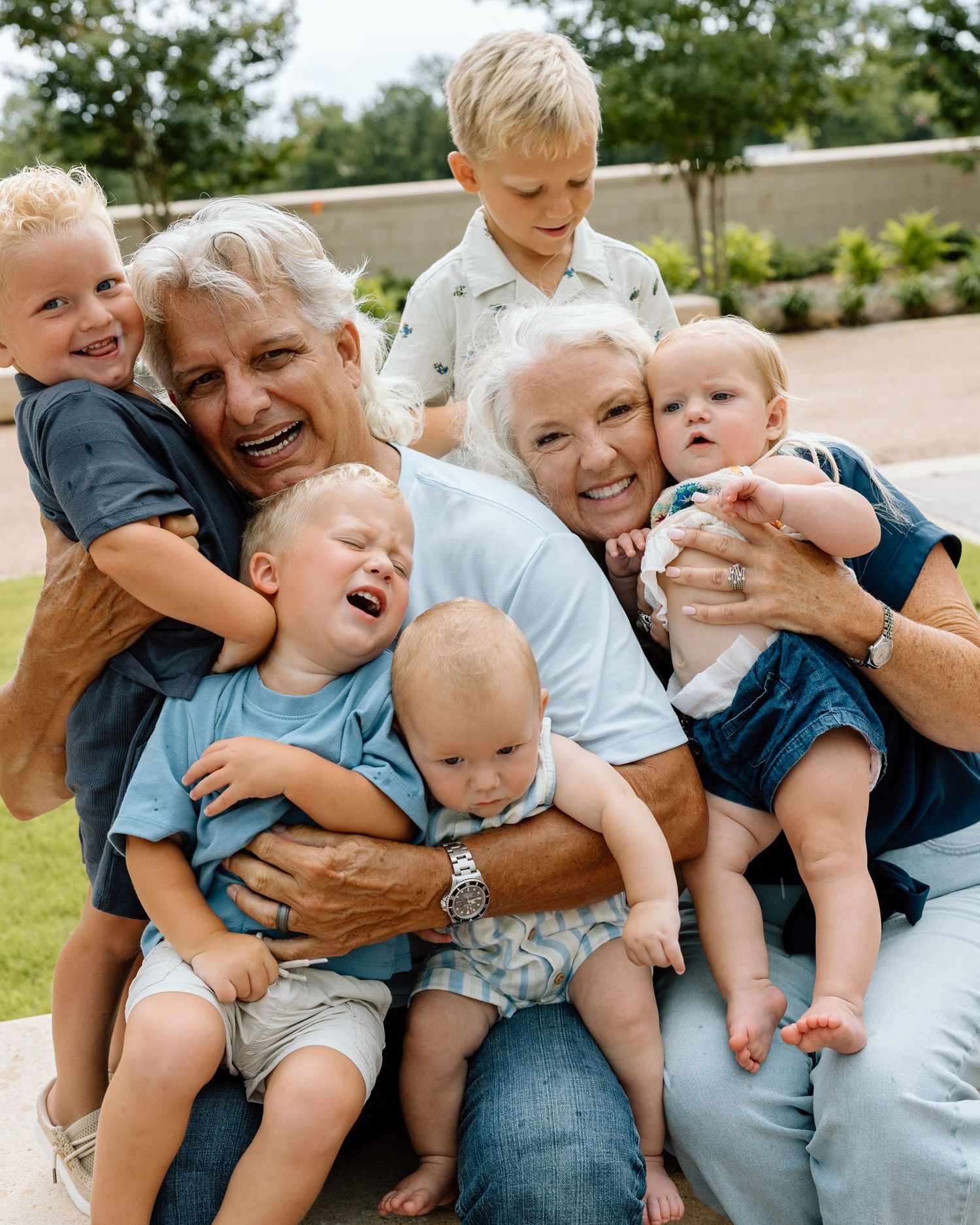 Grandparents joyfully surrounded by four young grandchildren in a warm outdoor family embrace.
