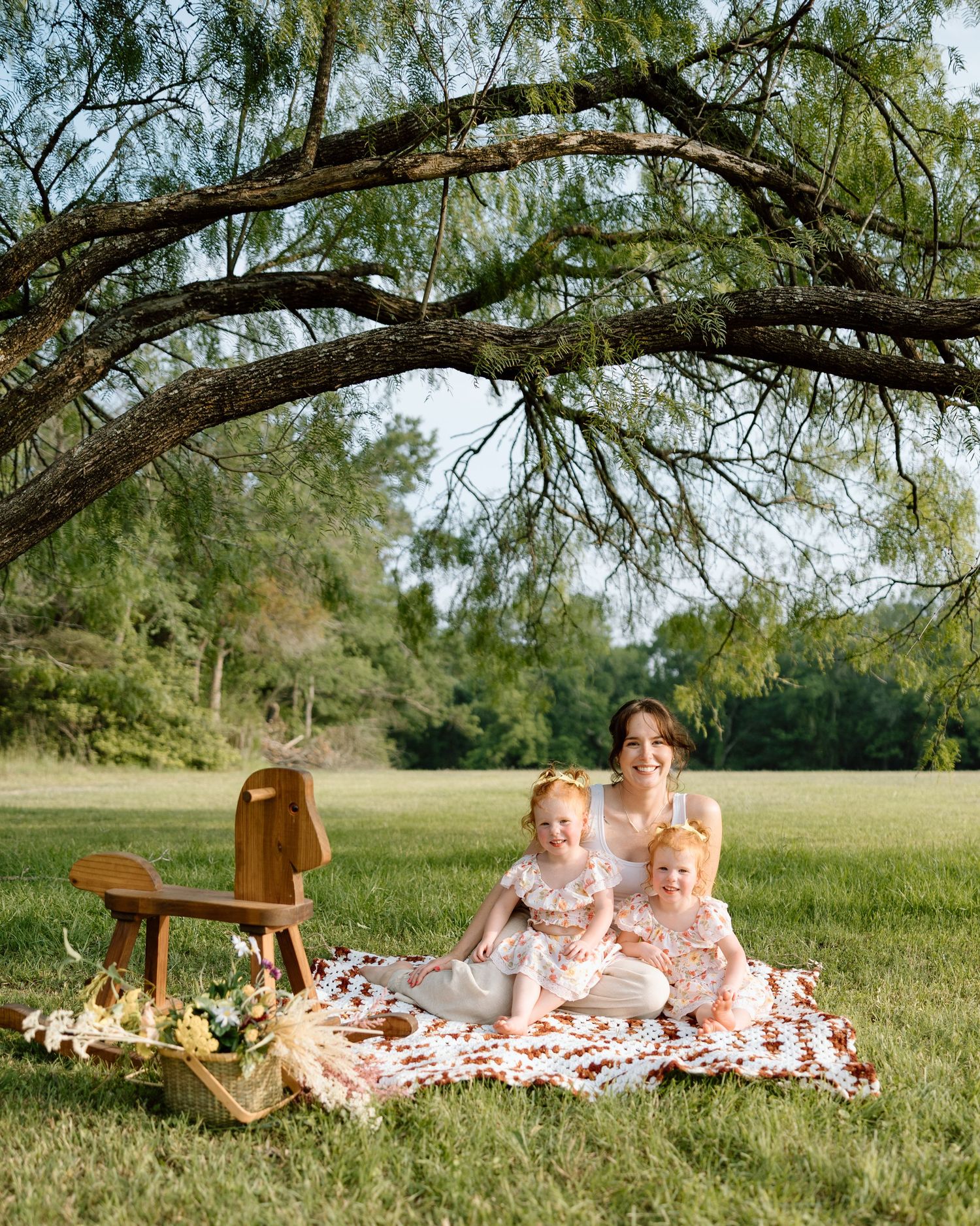Mother and two toddler girls sitting on a quilt under a large tree in a green meadow for a family picnic photo.