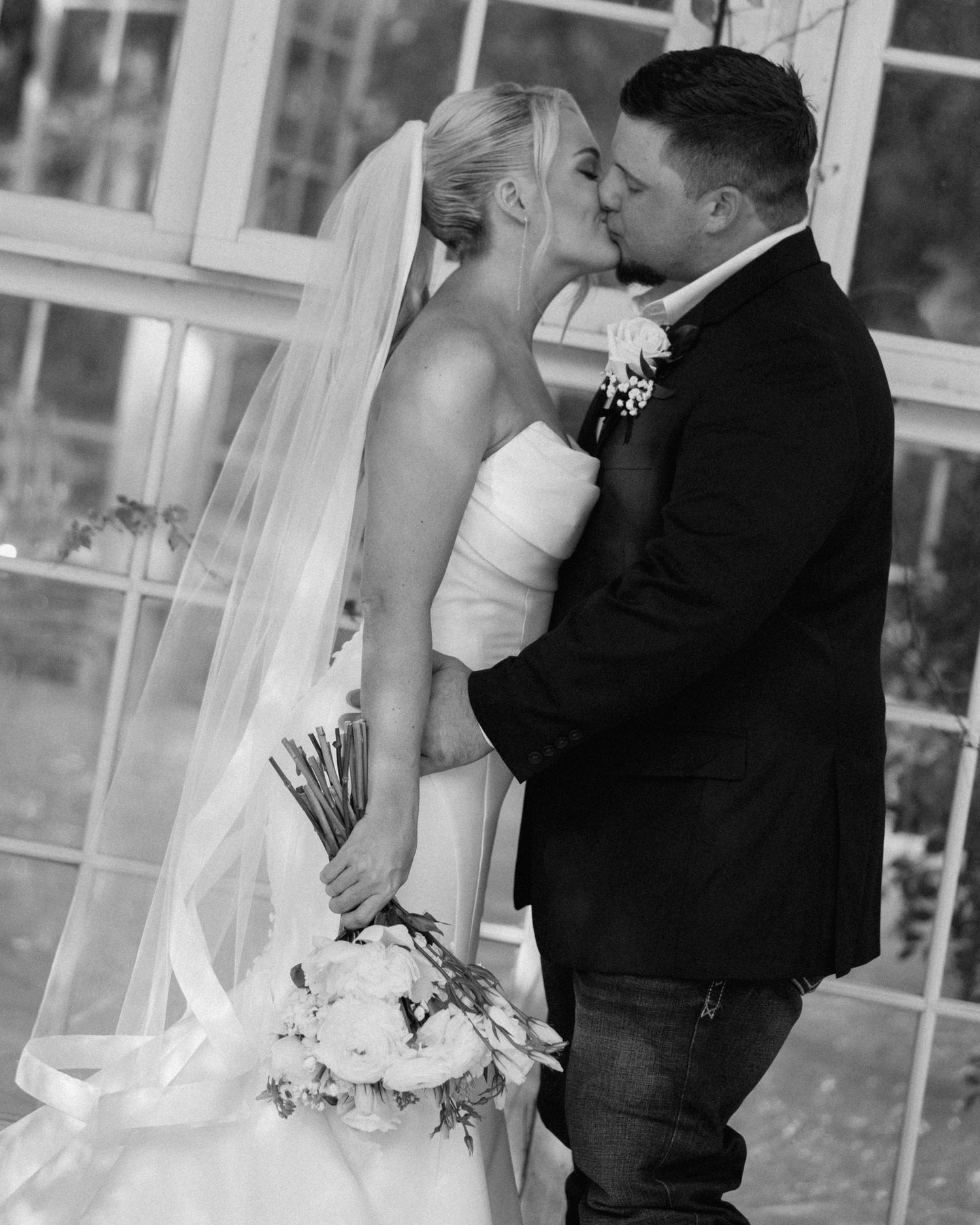 Black and white photo of bride and groom sharing first kiss, bride holding white bouquet before greenhouse windows.