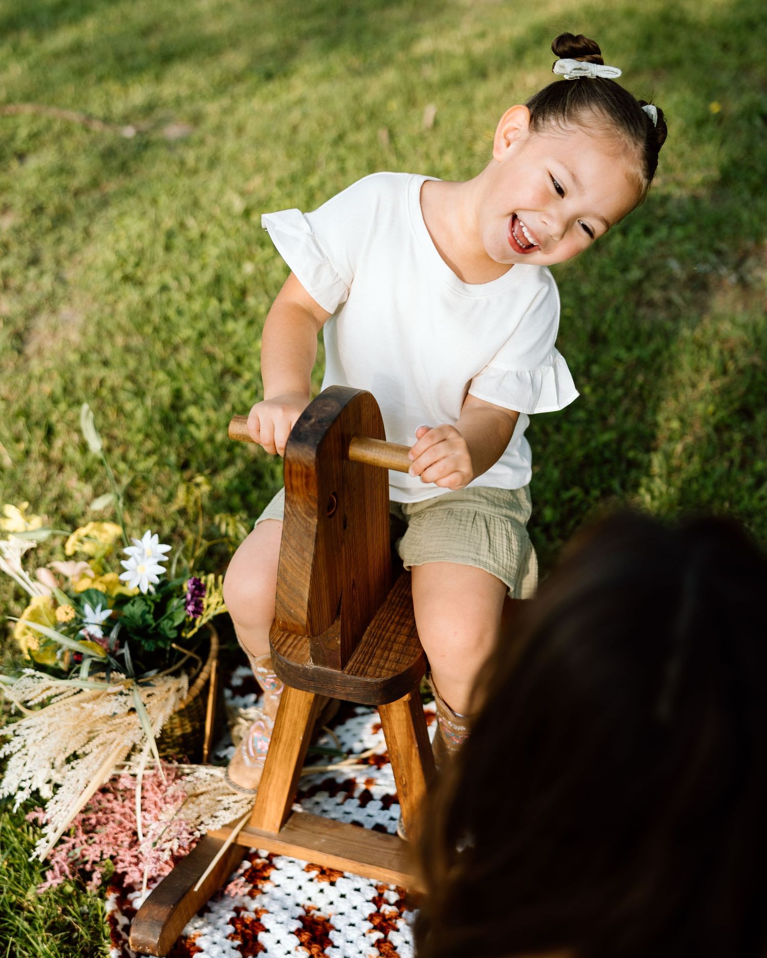 A joyful young girl plays on a wooden rocking horse toy outdoors on a crochet blanket in a grassy park.