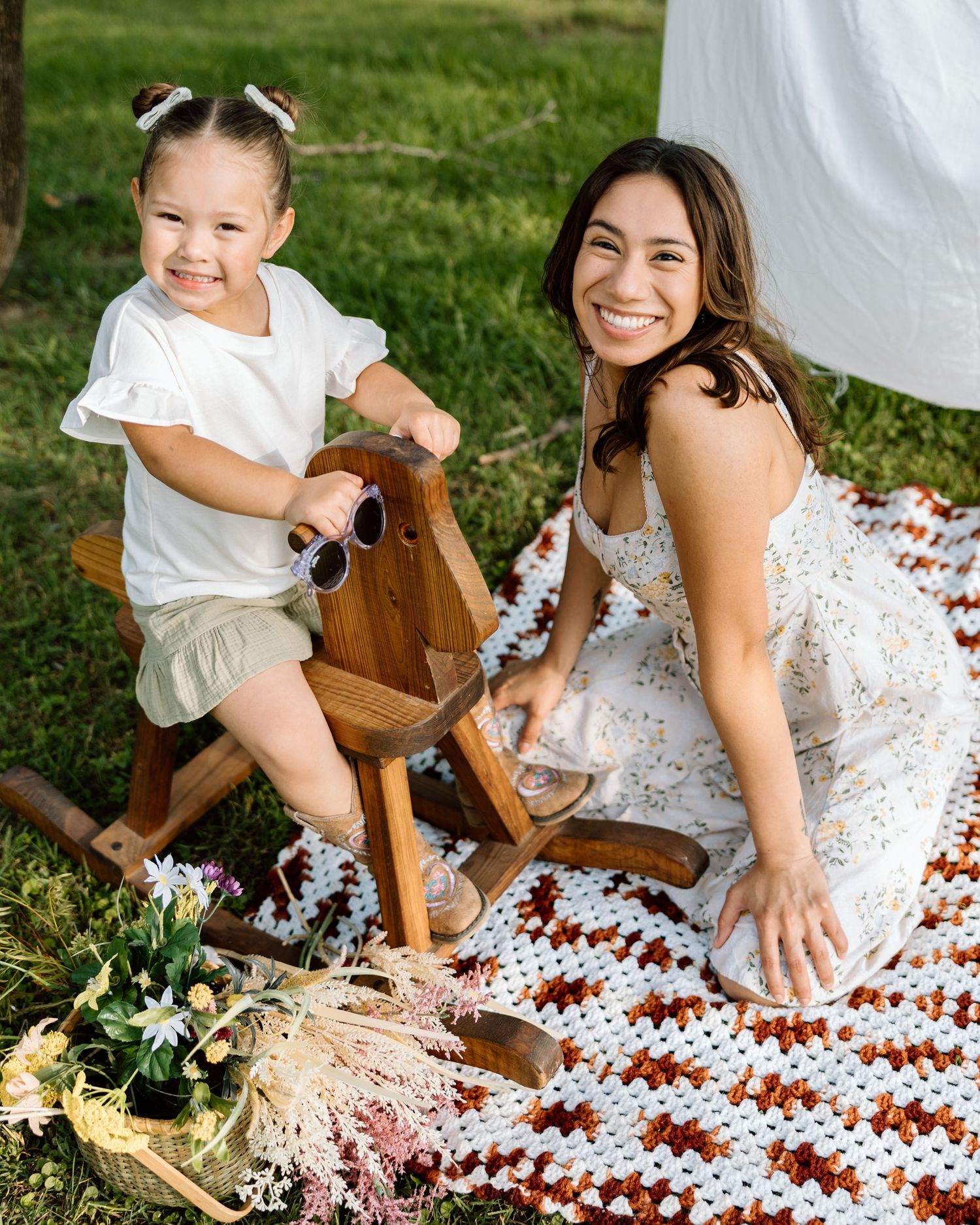 A smiling woman and toddler girl enjoy a sunny outdoor picnic on a crochet blanket with a wooden rocking horse.