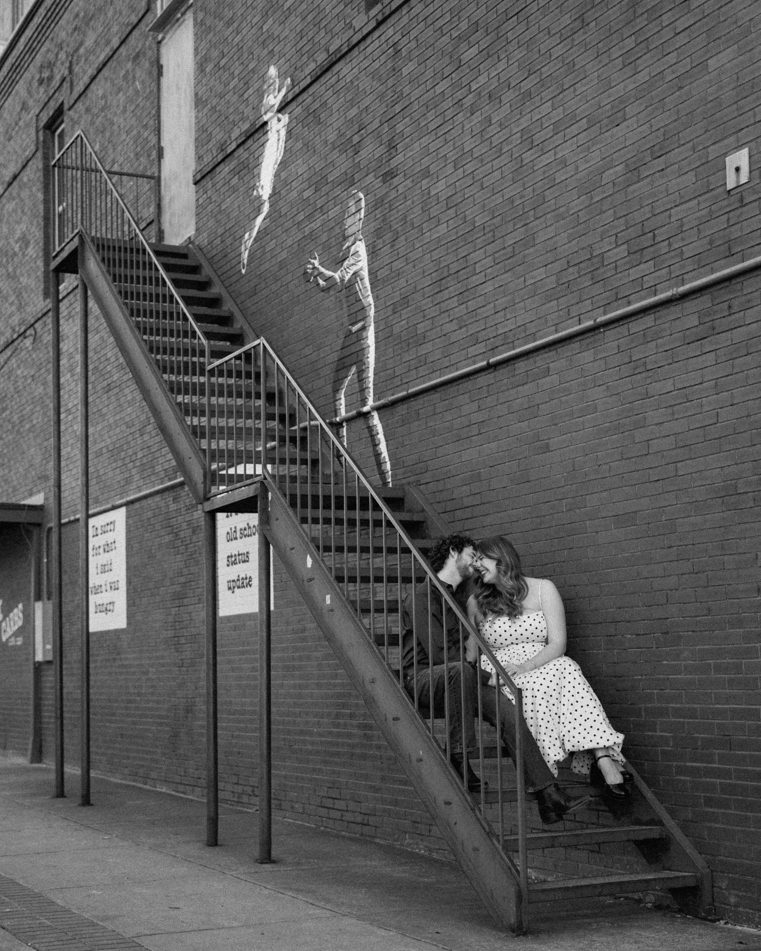 Black and white photo of a couple kissing on a metal fire escape staircase against a brick wall with street art.