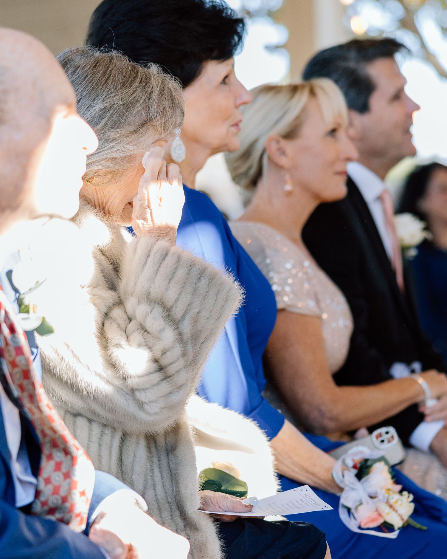 Emotional wedding guest wiping tears during outdoor ceremony, wearing blue dress and fur wrap.