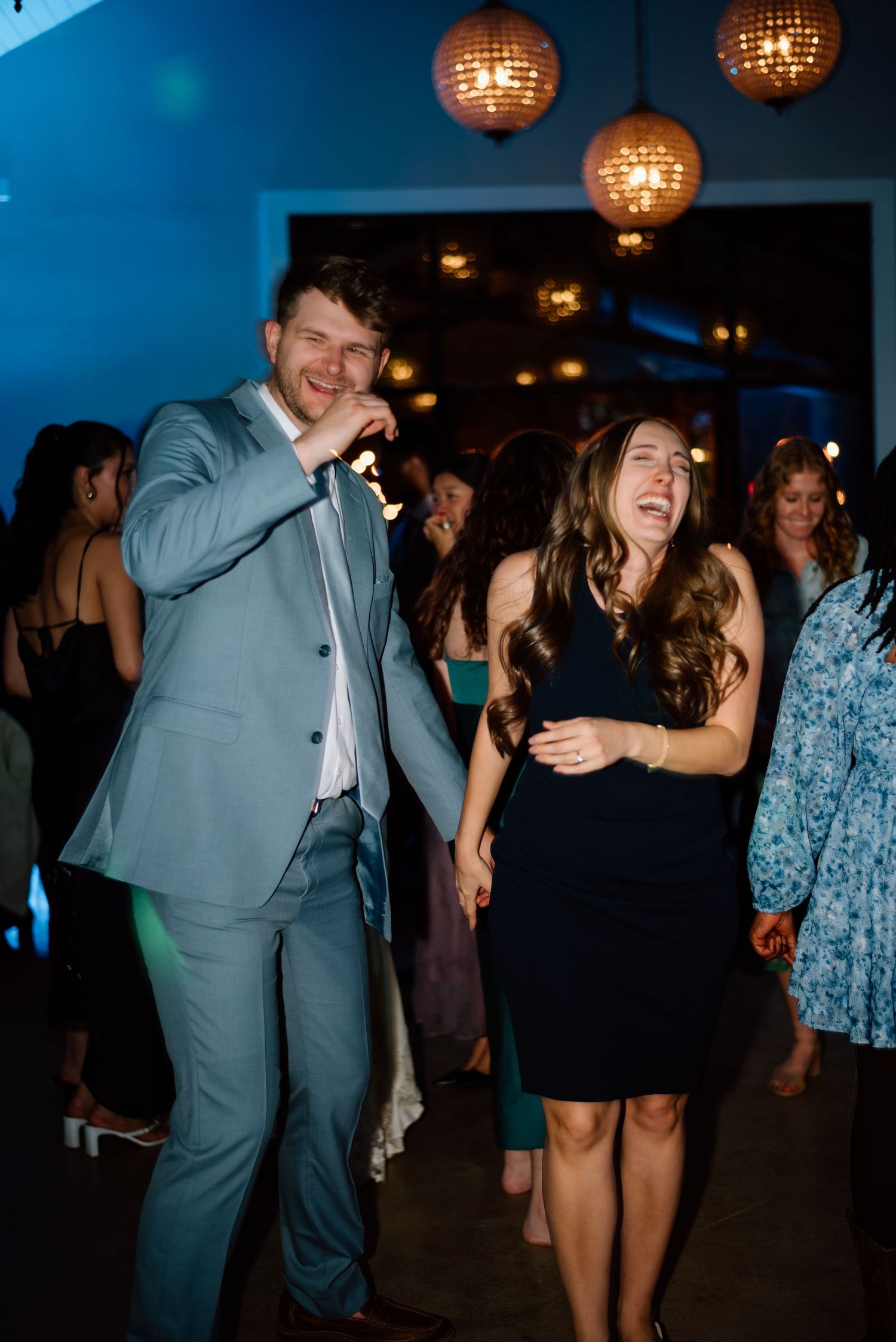 A man in a gray suit and woman in black dress laugh and dance together at a wedding reception.