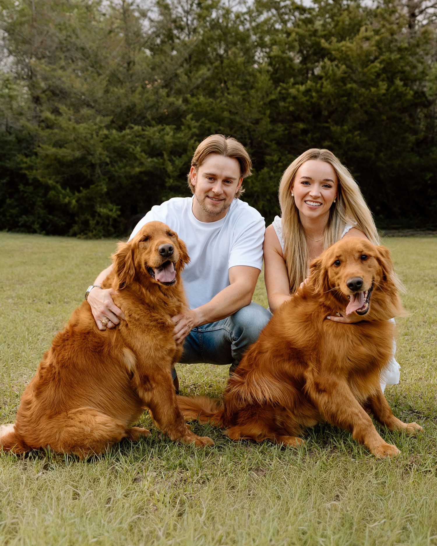 A smiling couple poses outdoors on green grass with two golden retrievers, surrounded by lush trees.