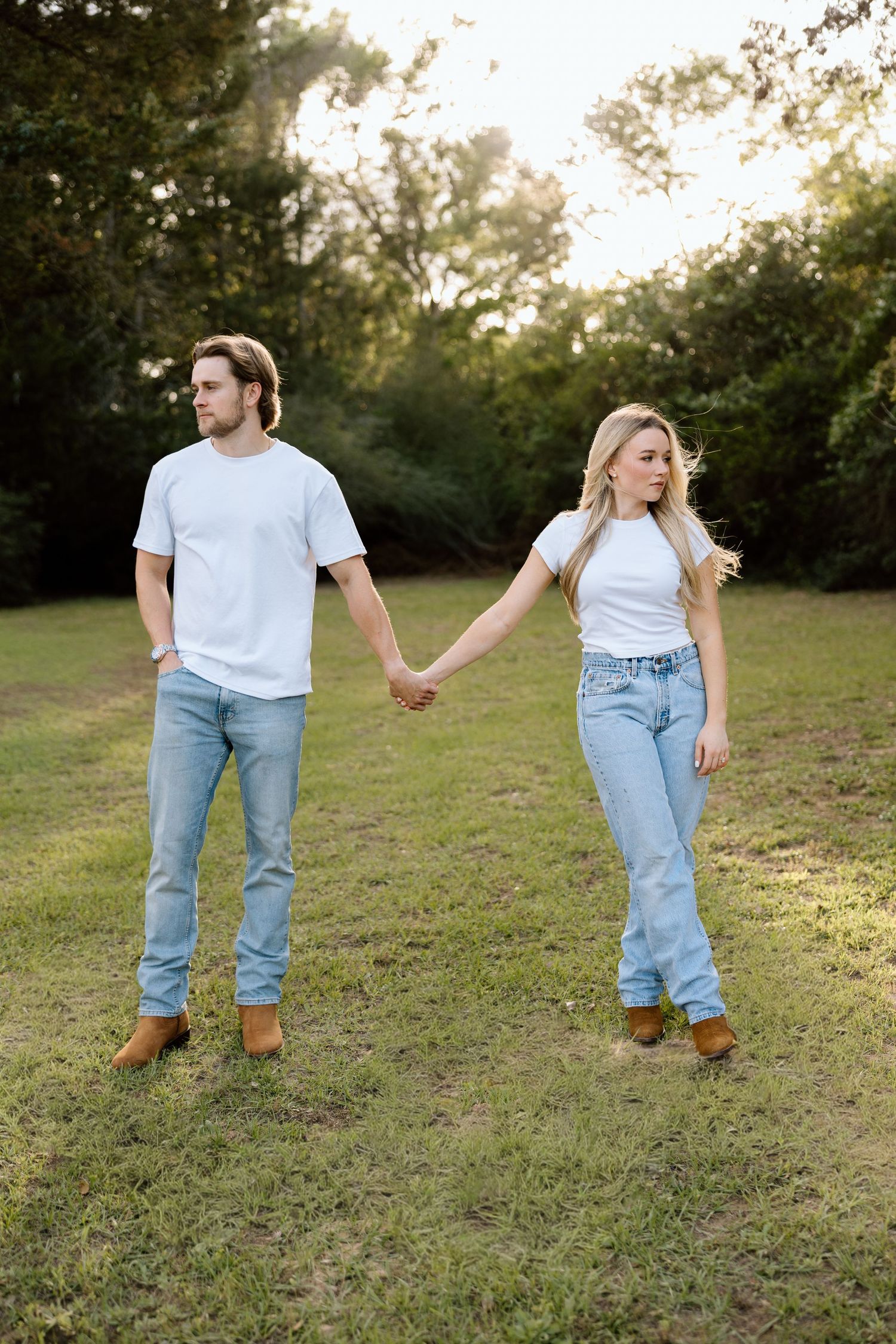 Engaged couple holding hands in a green park wearing matching white tees and light blue jeans.