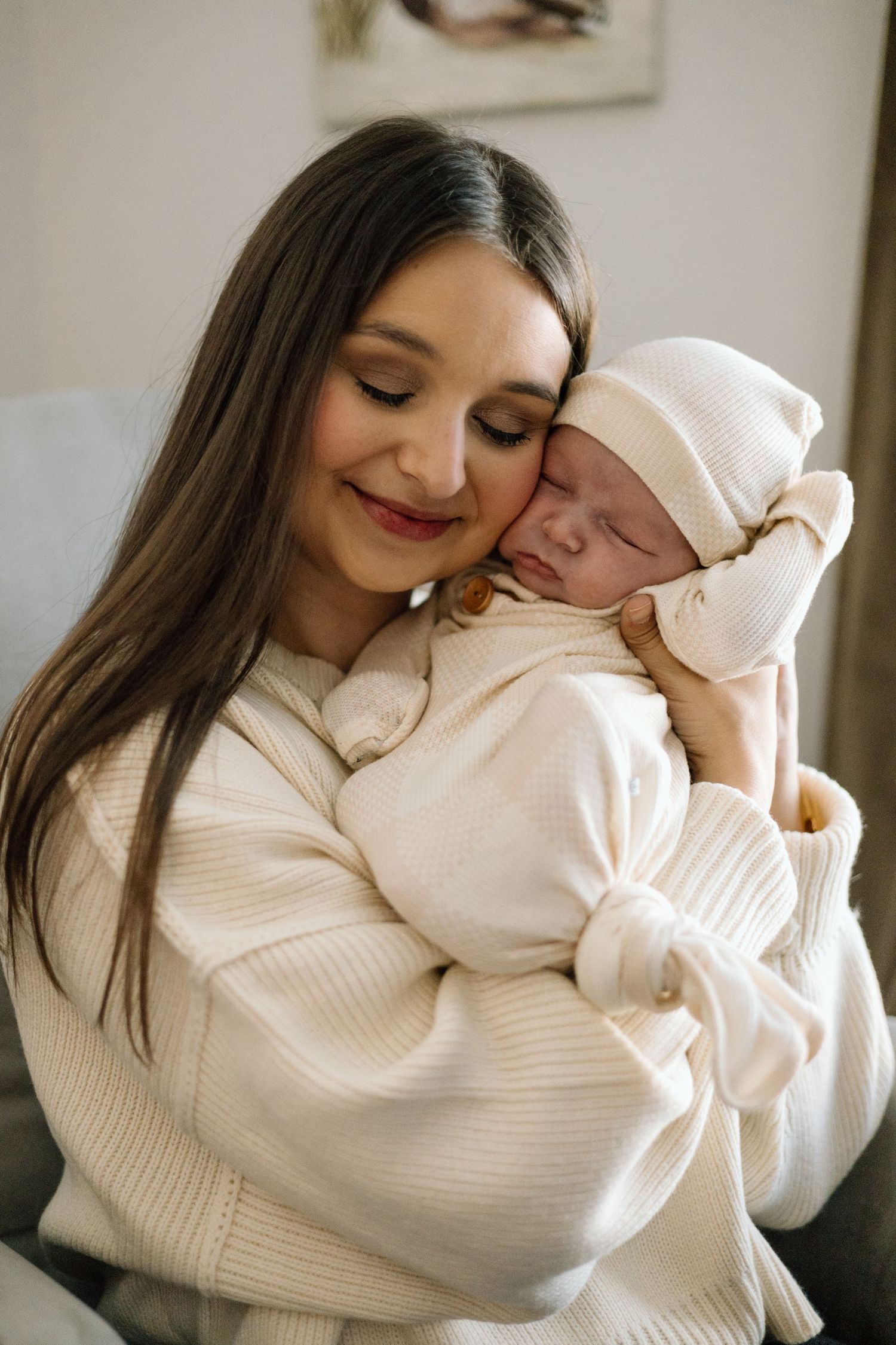 Smiling mother in cream sweater tenderly holds sleeping newborn baby dressed in white knit outfit and hat.
