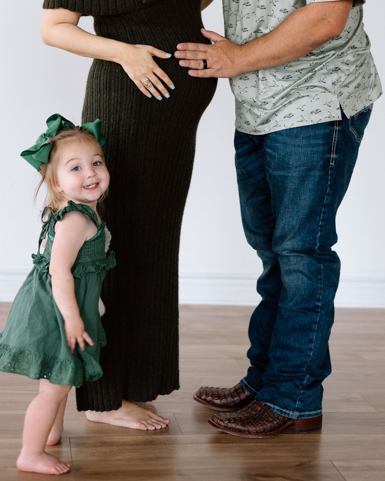 A toddler girl in a green dress smiles beside her pregnant mother and father during a maternity photo session.