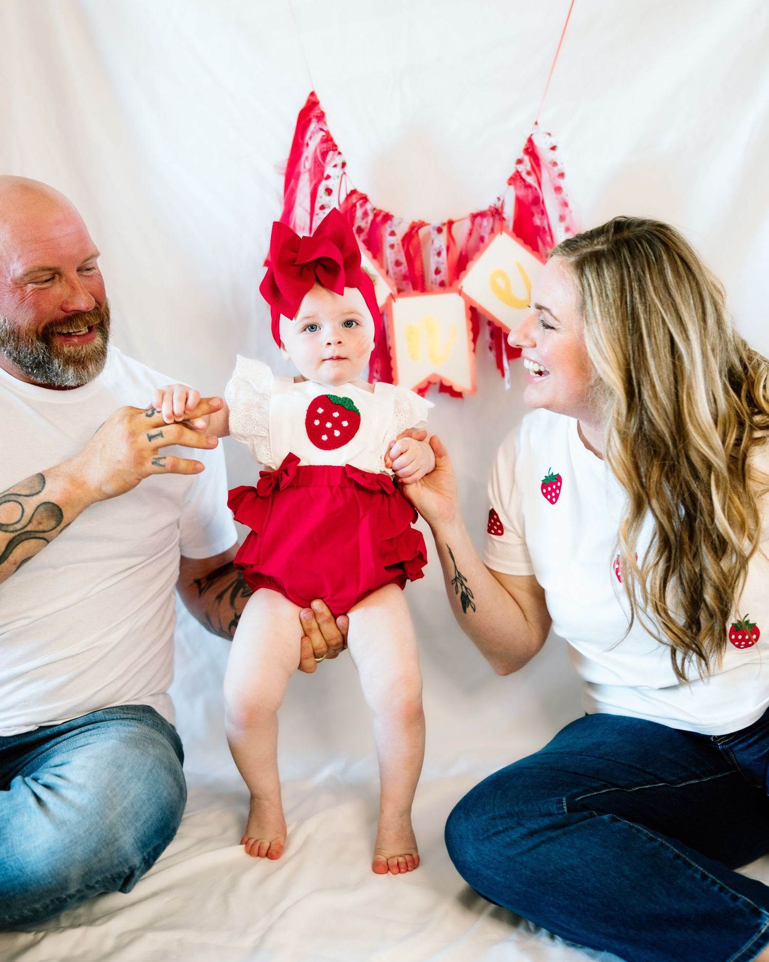 Family photo with parents holding baby girl in red strawberry outfit and bow headband for first birthday celebration.
