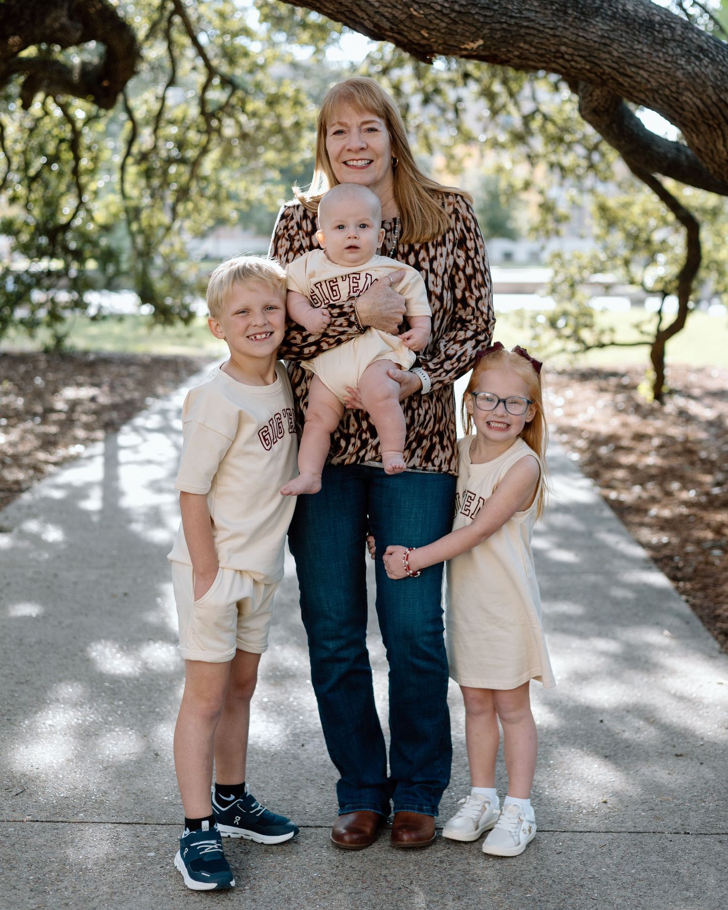 A smiling mother holds a baby while posing outdoors with two young children wearing matching cream outfits.