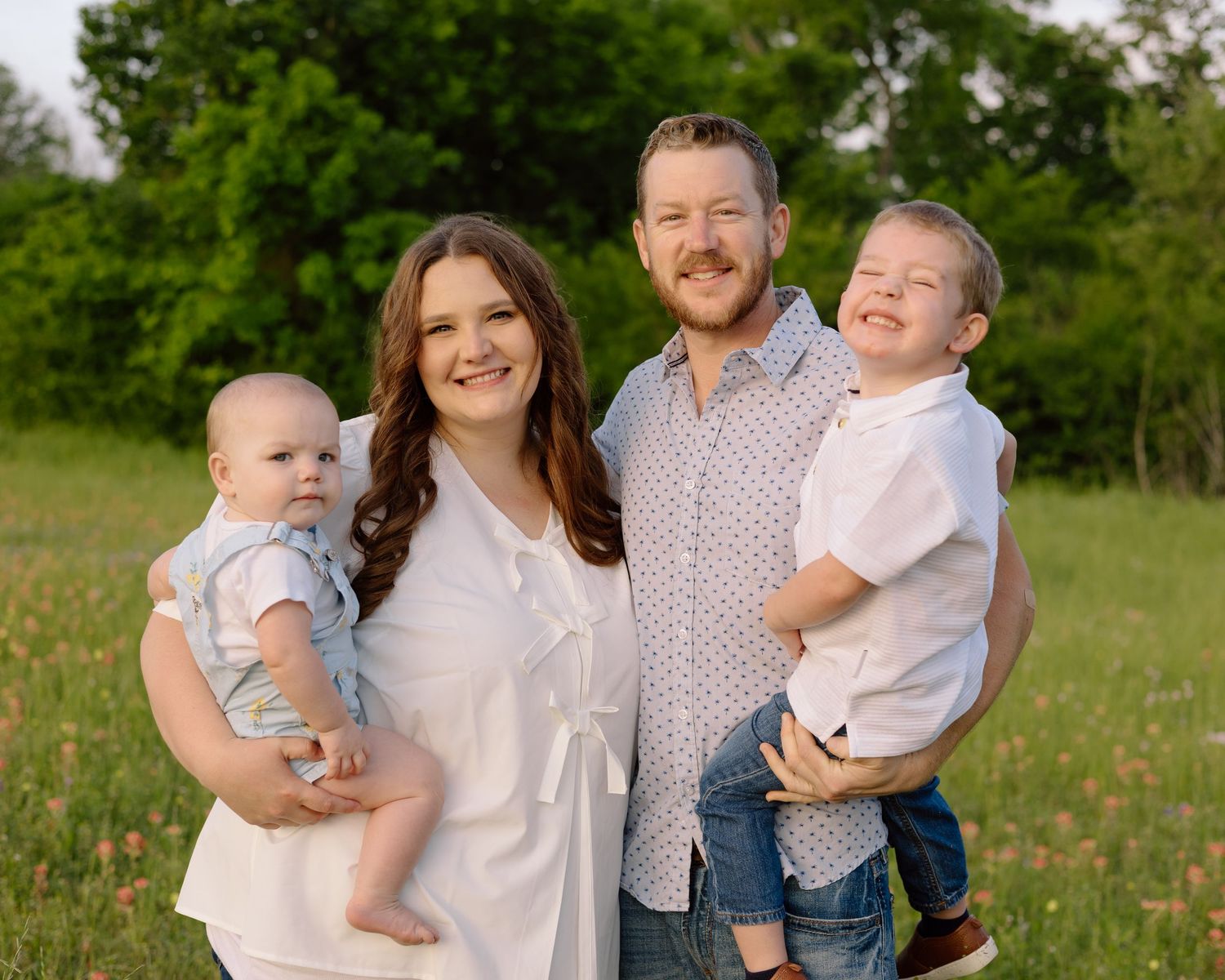 Family of four outdoors in a sunny meadow with green trees in the background.