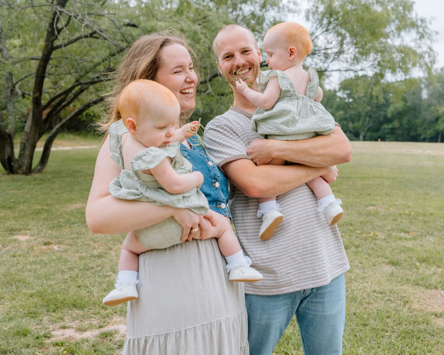 A couple stands outdoors in a park setting holding two babies wearing matching outfits on a sunny day.