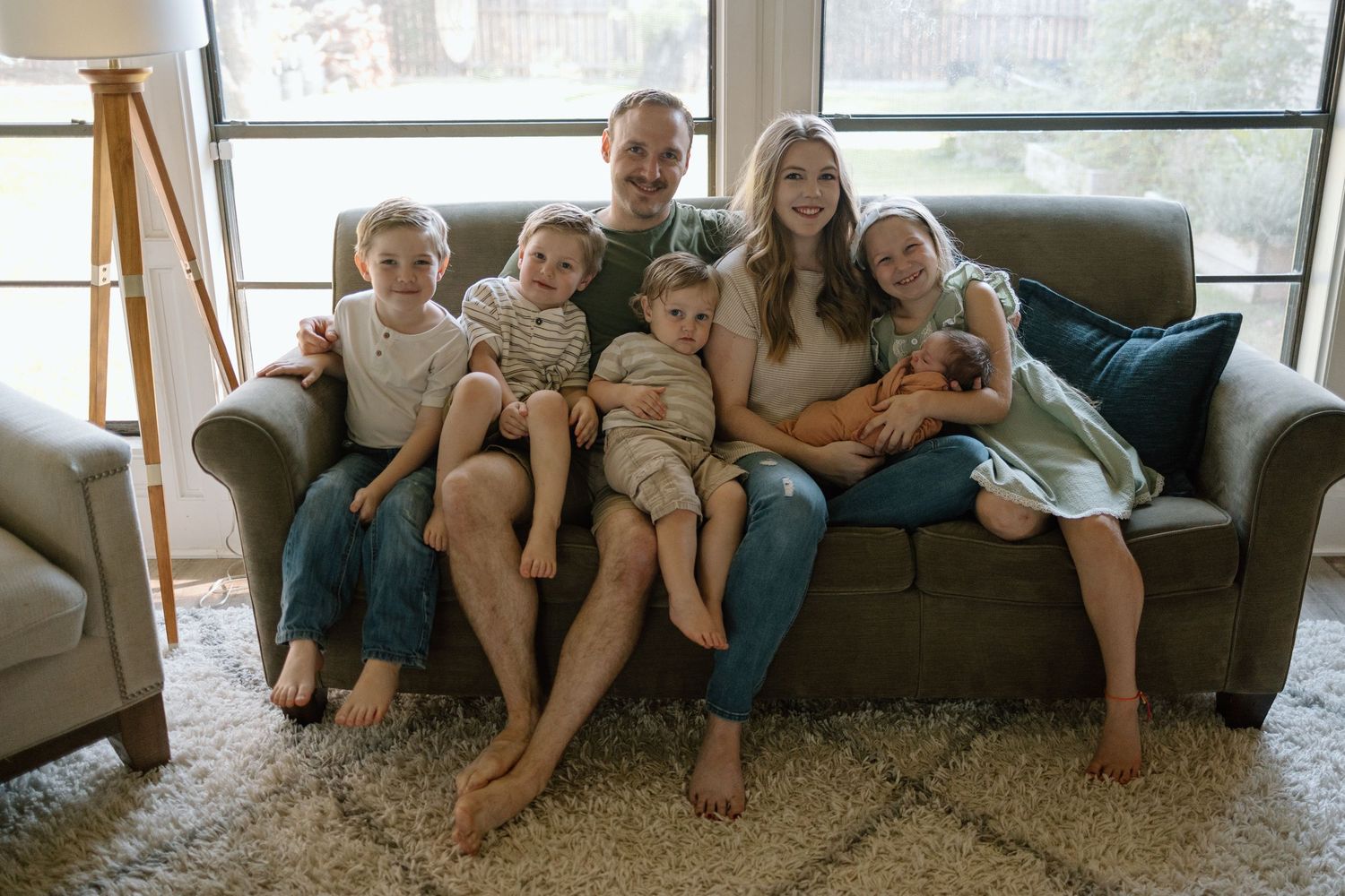 A group sits close together on a gray couch in a sunlit living room with a plush area rug.