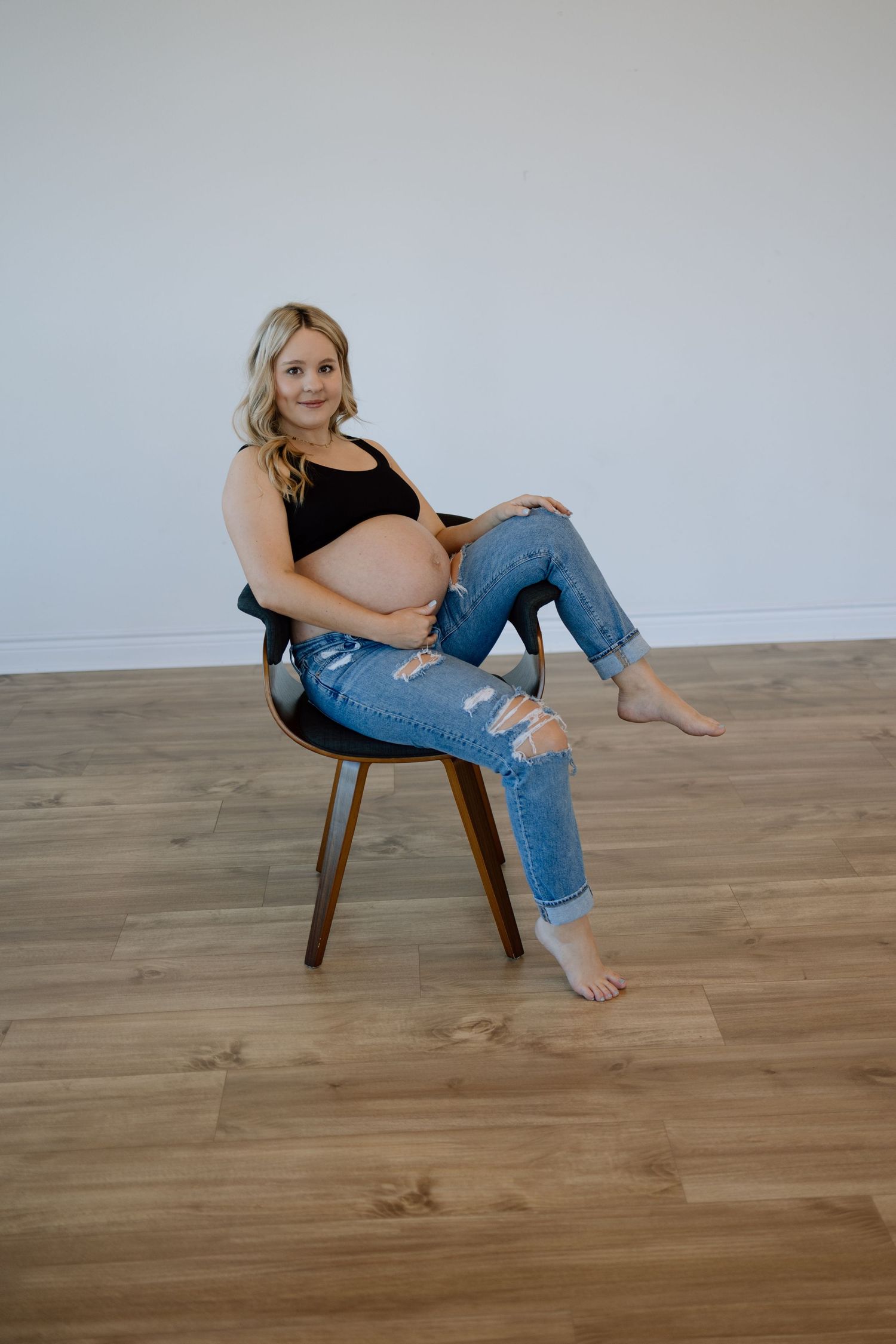 Person wearing ripped jeans and black top sits casually in wooden chair on hardwood floor in studio setting.