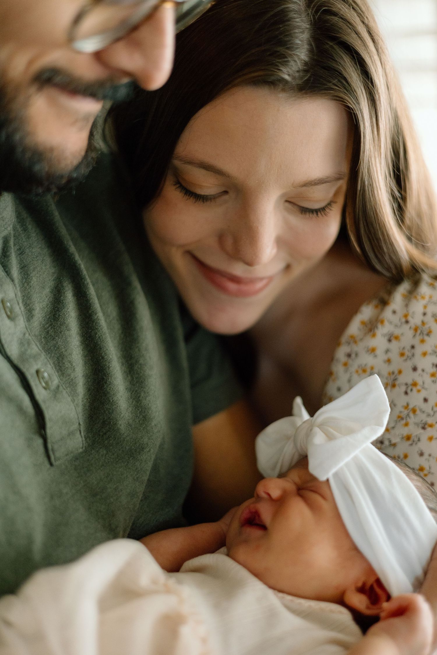 Intimate close-up of parents admiring their sleeping newborn with white bow headband.