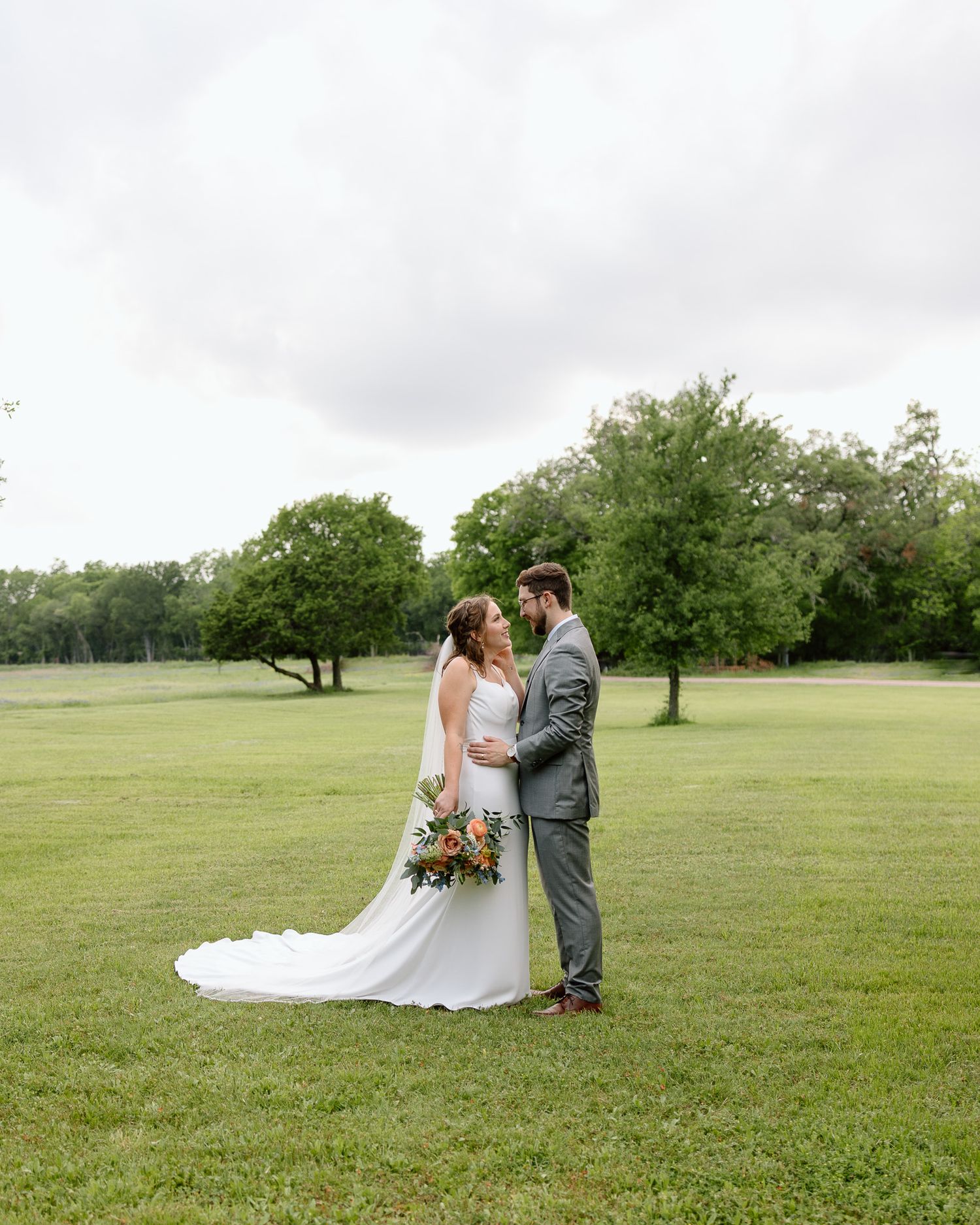 Newlyweds share intimate moment on expansive green lawn with cloudy sky backdrop.